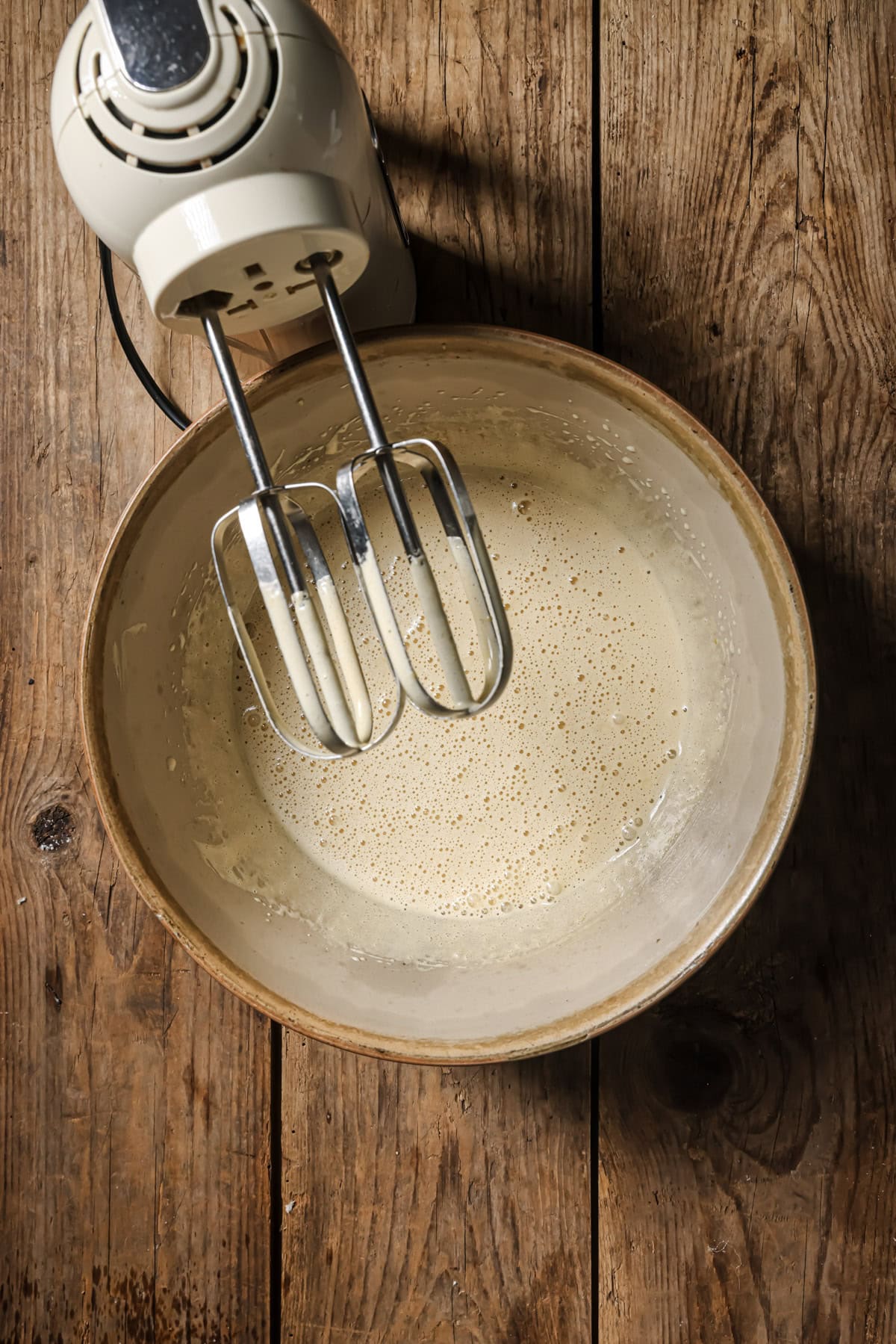 An electric hand mixer with beaters rests over a mixing bowl filled with a pale, frothy batter on a rustic wooden surface.