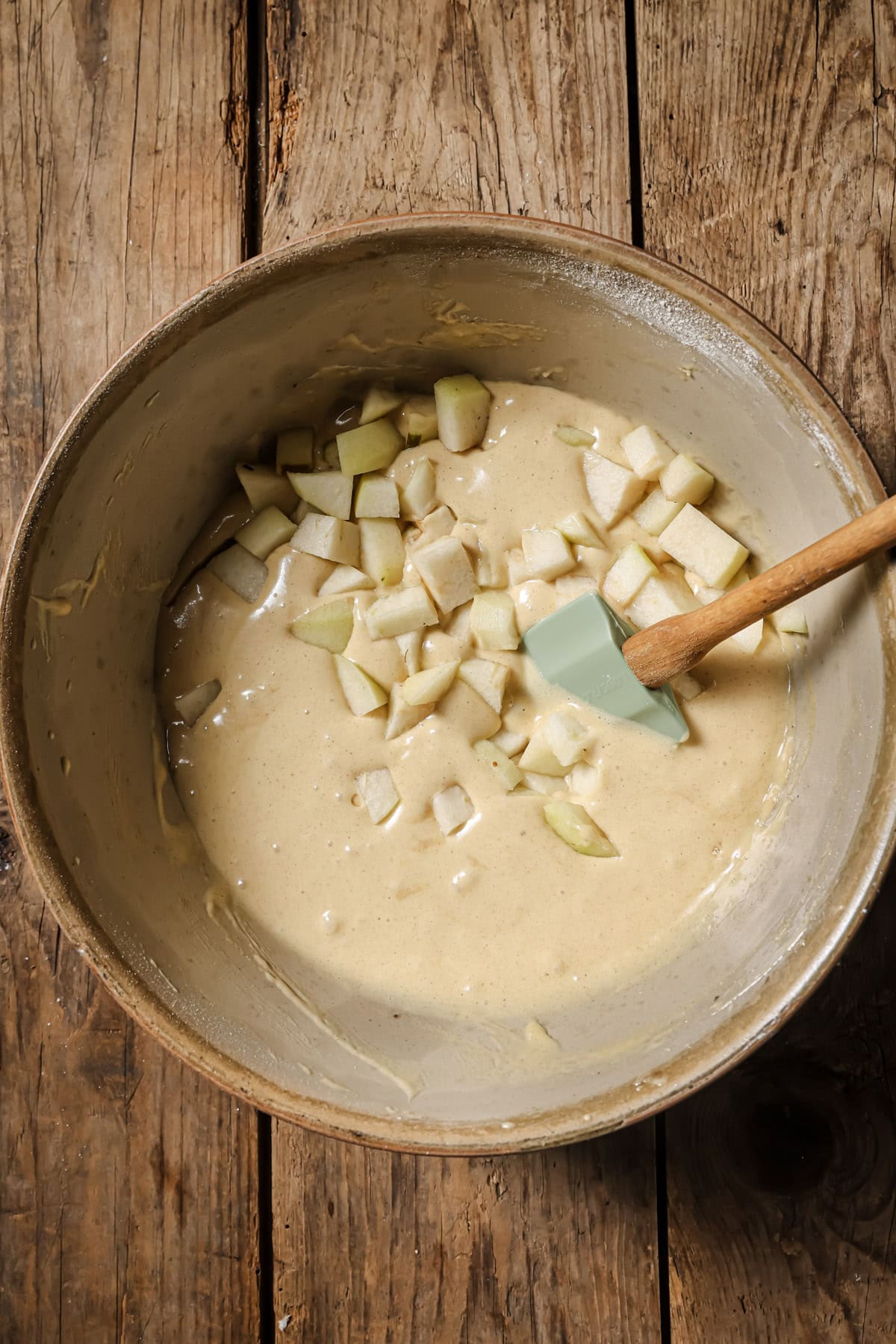 A mixing bowl with cake batter and chopped apples, being stirred with a spatula, sits on a rustic wooden surface.