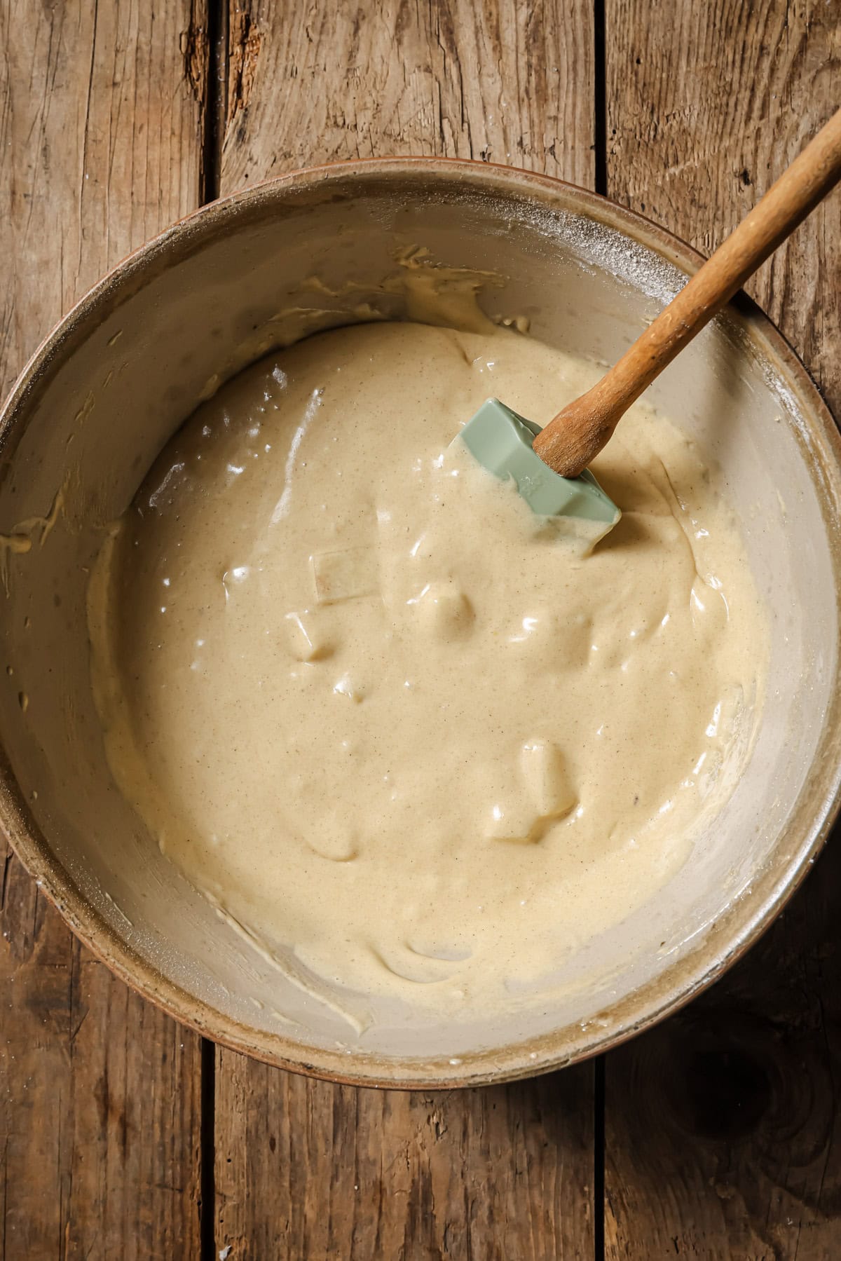 A bowl of thick, creamy batter with visible chunks is being mixed with a silicone spatula on a rustic wooden surface.