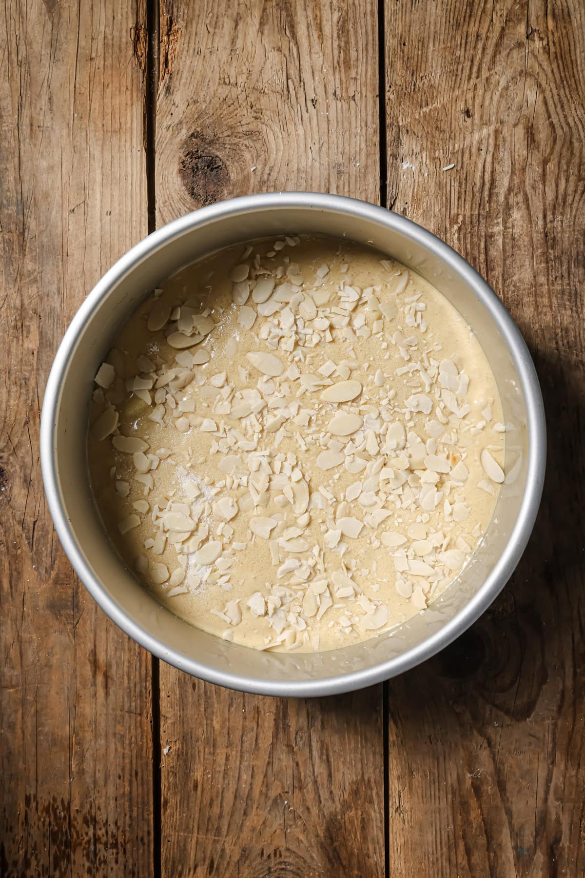 A round metal cake pan filled with unbaked cake batter topped with sliced almonds, sitting on a rustic wooden surface.