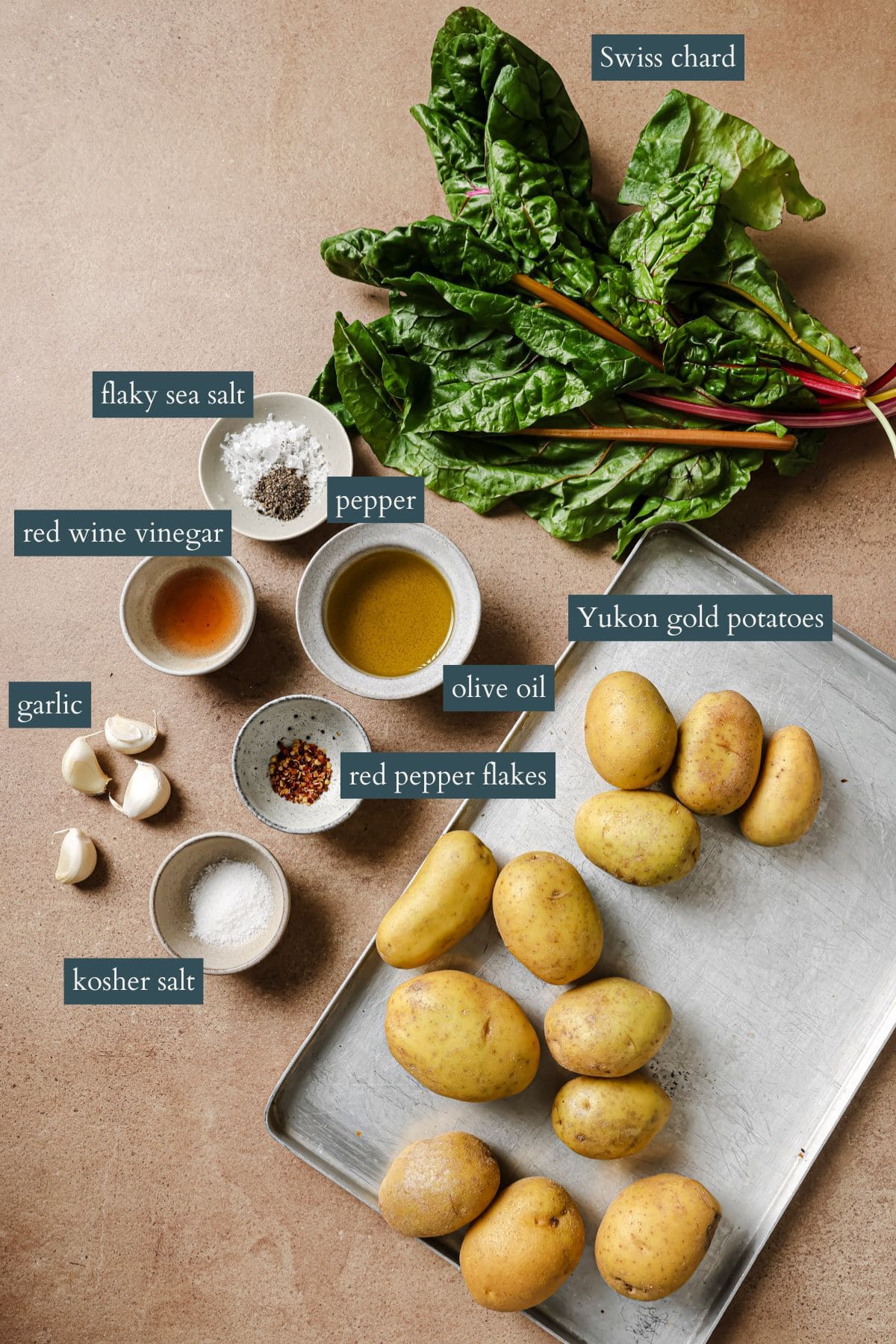 Ingredients for a recipe are arranged on a surface, including Yukon gold potatoes on a tray, Swiss chard, bowls of kosher and flaky sea salt, pepper, red wine vinegar, olive oil, red pepper flakes, and garlic cloves.
