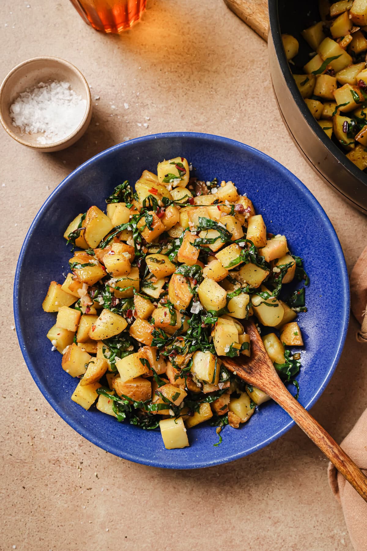 A blue bowl filled with sautéed diced potatoes, wilted greens, and herbs, with a wooden spoon resting inside. A small bowl of salt and a skillet with more of the dish are nearby on a tan surface.