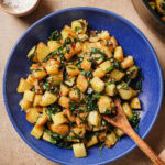 A blue bowl filled with sautéed diced potatoes and greens, garnished with herbs, sits on a beige surface. A wooden spoon rests in the bowl, and a small dish of salt is nearby.