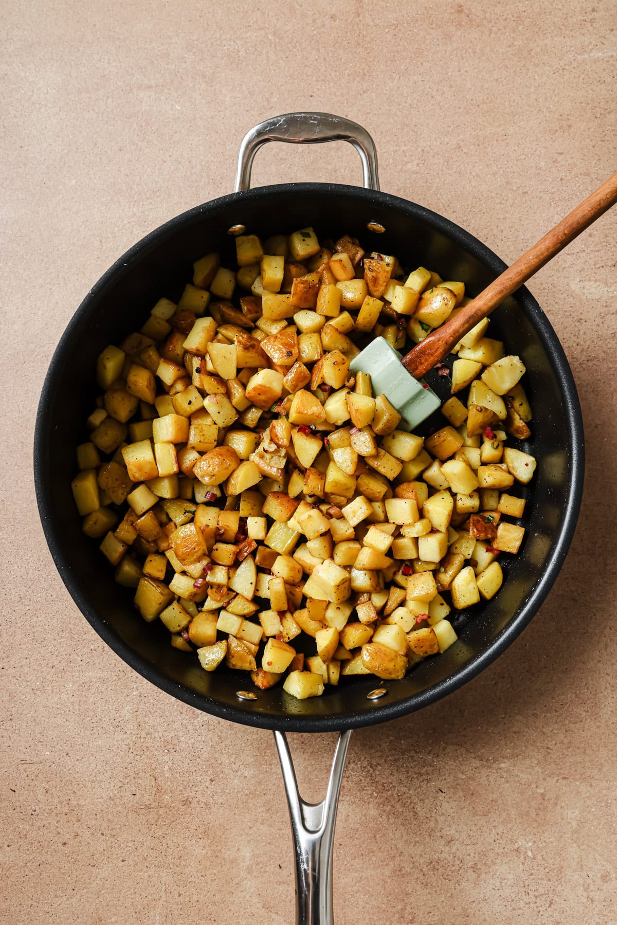 A skillet filled with diced, cooked potatoes seasoned with spices, with a wooden-handled spatula resting inside the pan, all on a light brown countertop.