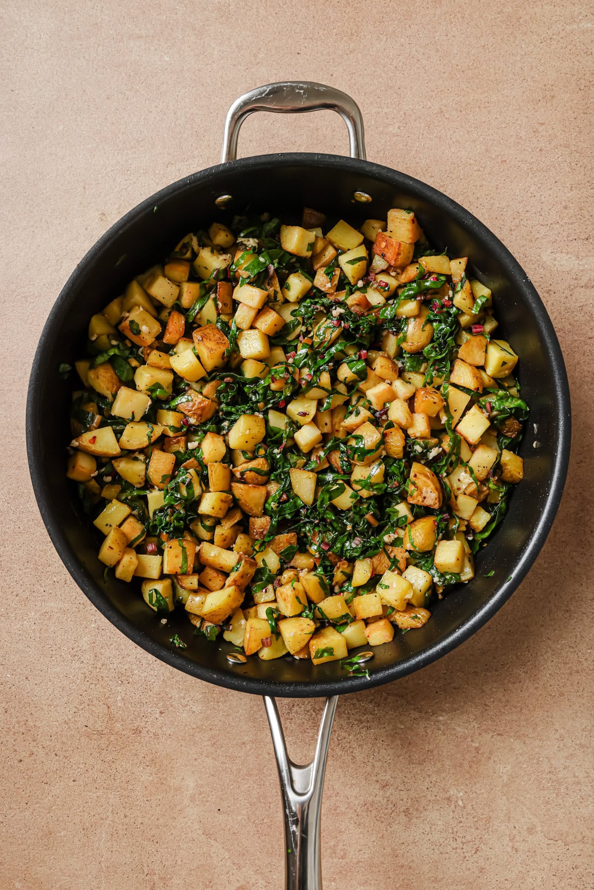 A skillet filled with sautéed diced potatoes, spinach, and herbs, sitting on a light brown countertop. The mixture appears golden and freshly cooked.