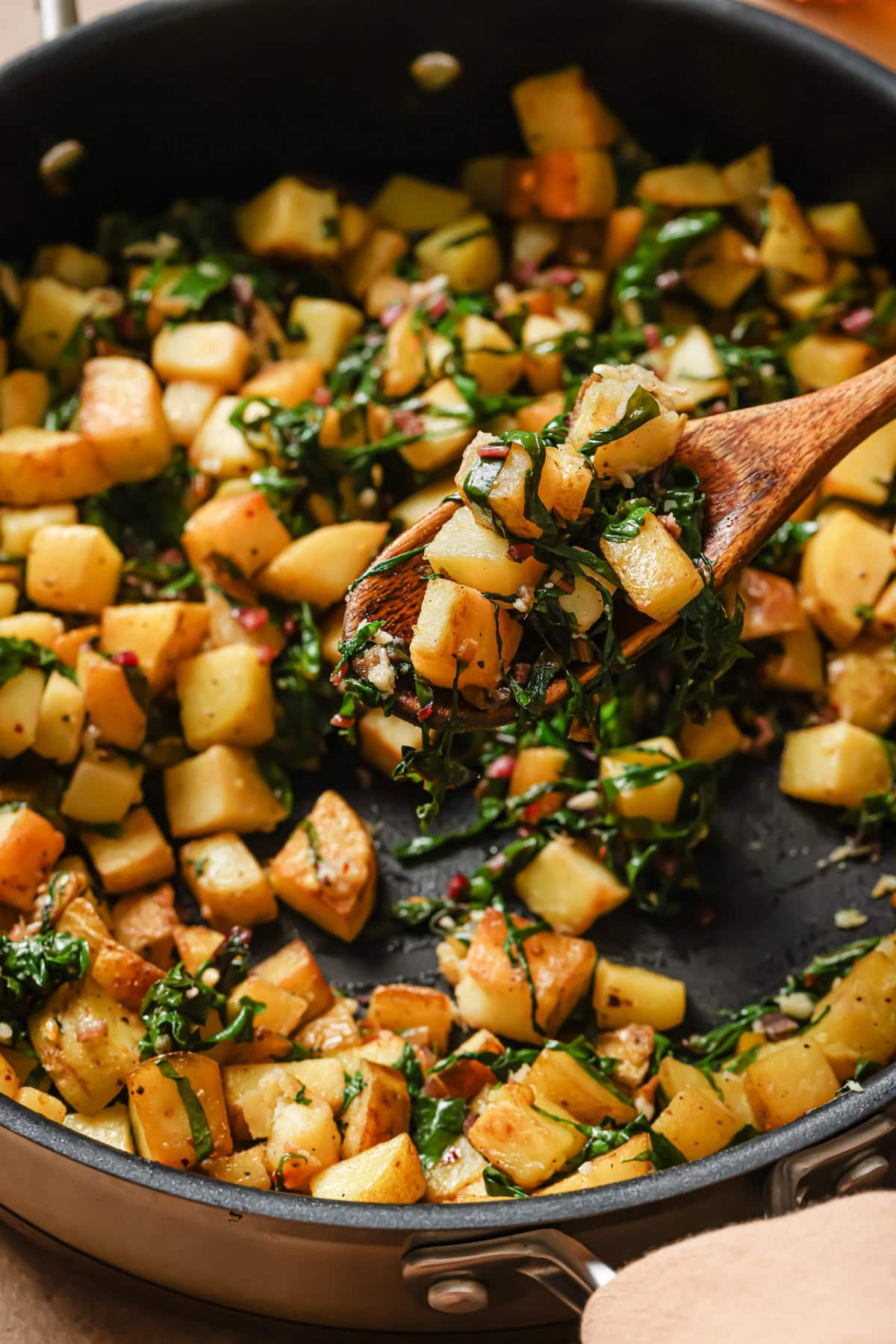 A skillet filled with sautéed diced potatoes and leafy greens, with a wooden spoon lifting a portion of the mixture. The vegetables are golden and lightly seasoned.
