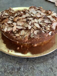 A round French pear cake topped with sliced almonds sits on a decorative gold plate. The cake has a golden-brown crust and appears moist, with some crumbs visible on the plate and countertop.