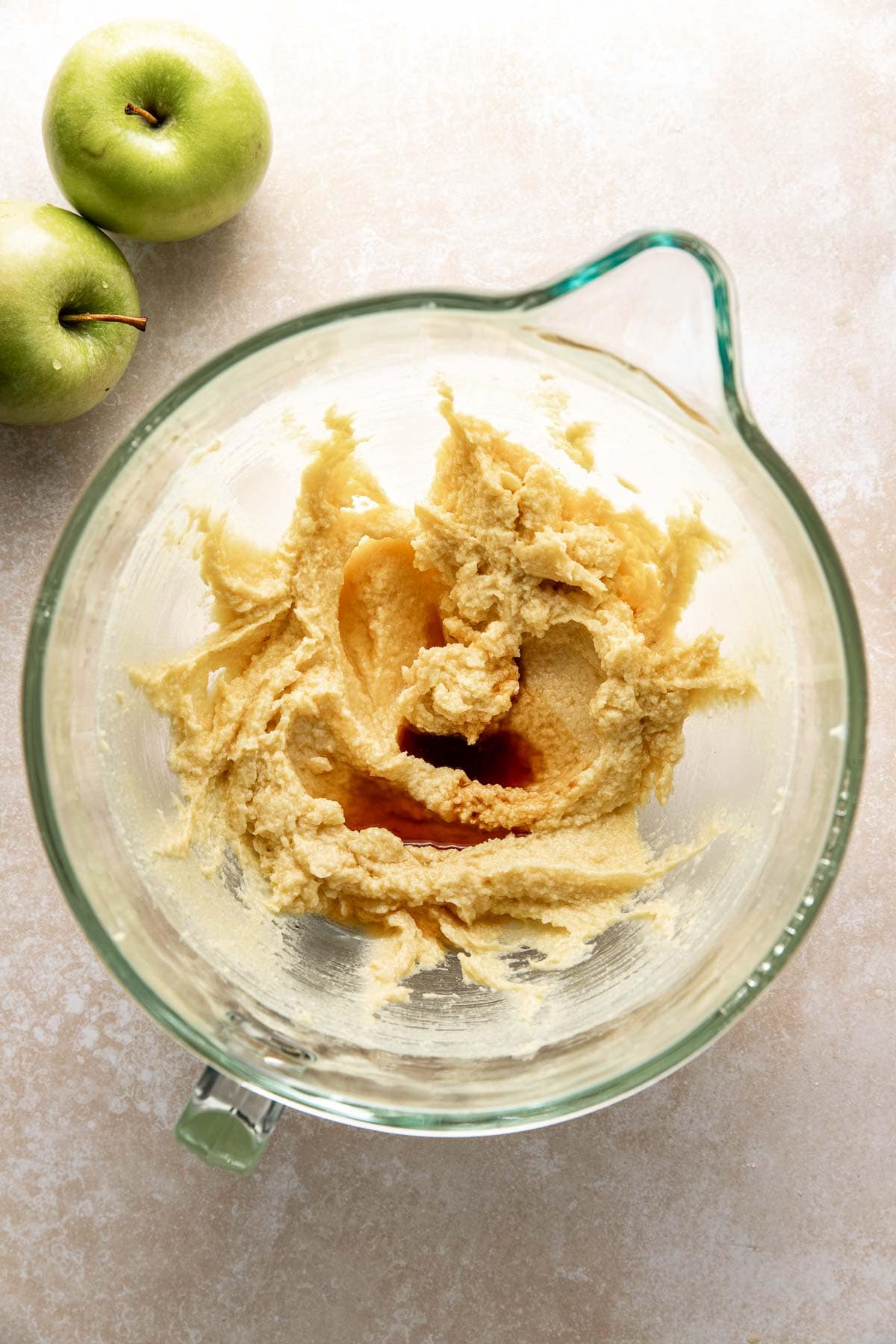 A glass mixing bowl with creamed butter and sugar sits on a light countertop next to two green apples.
