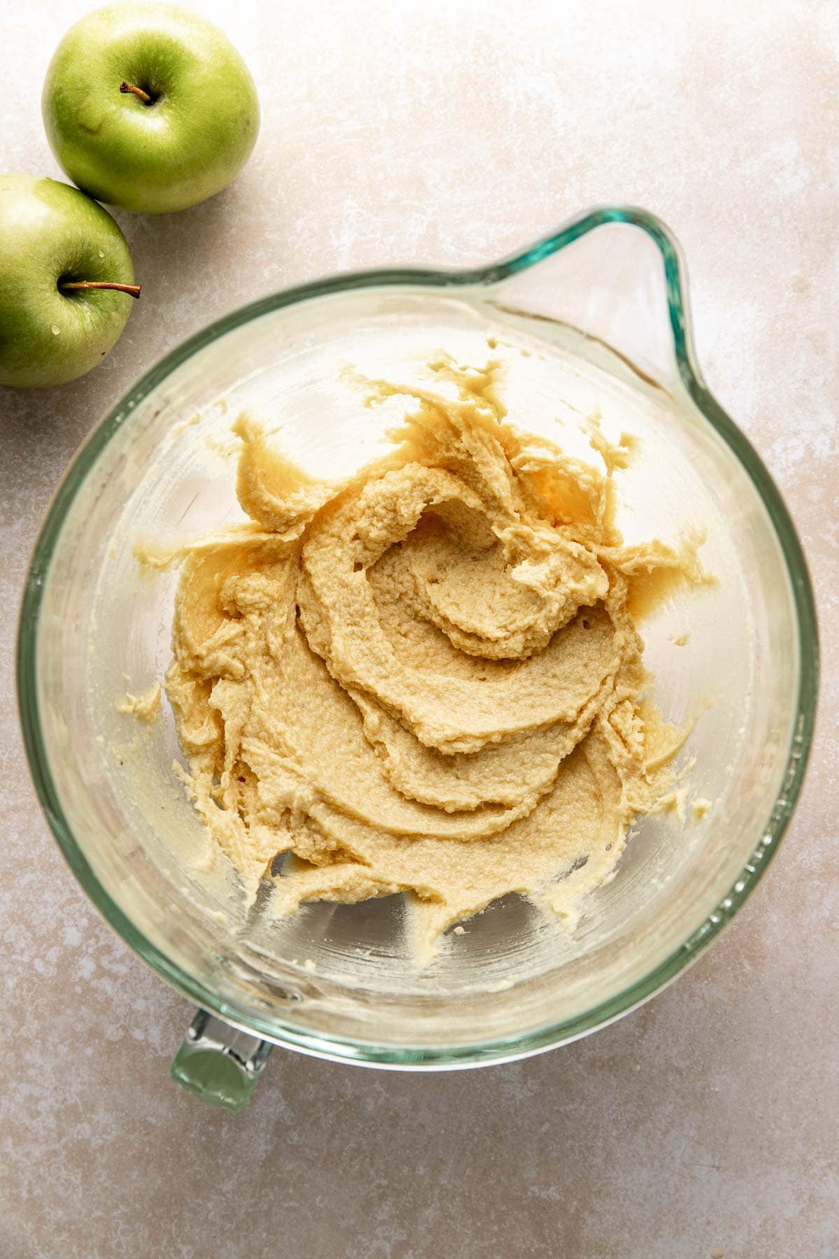A glass mixing bowl filled with creamy, light brown batter sits on a light-colored surface. Two green apples are placed nearby in the upper left corner of the image.