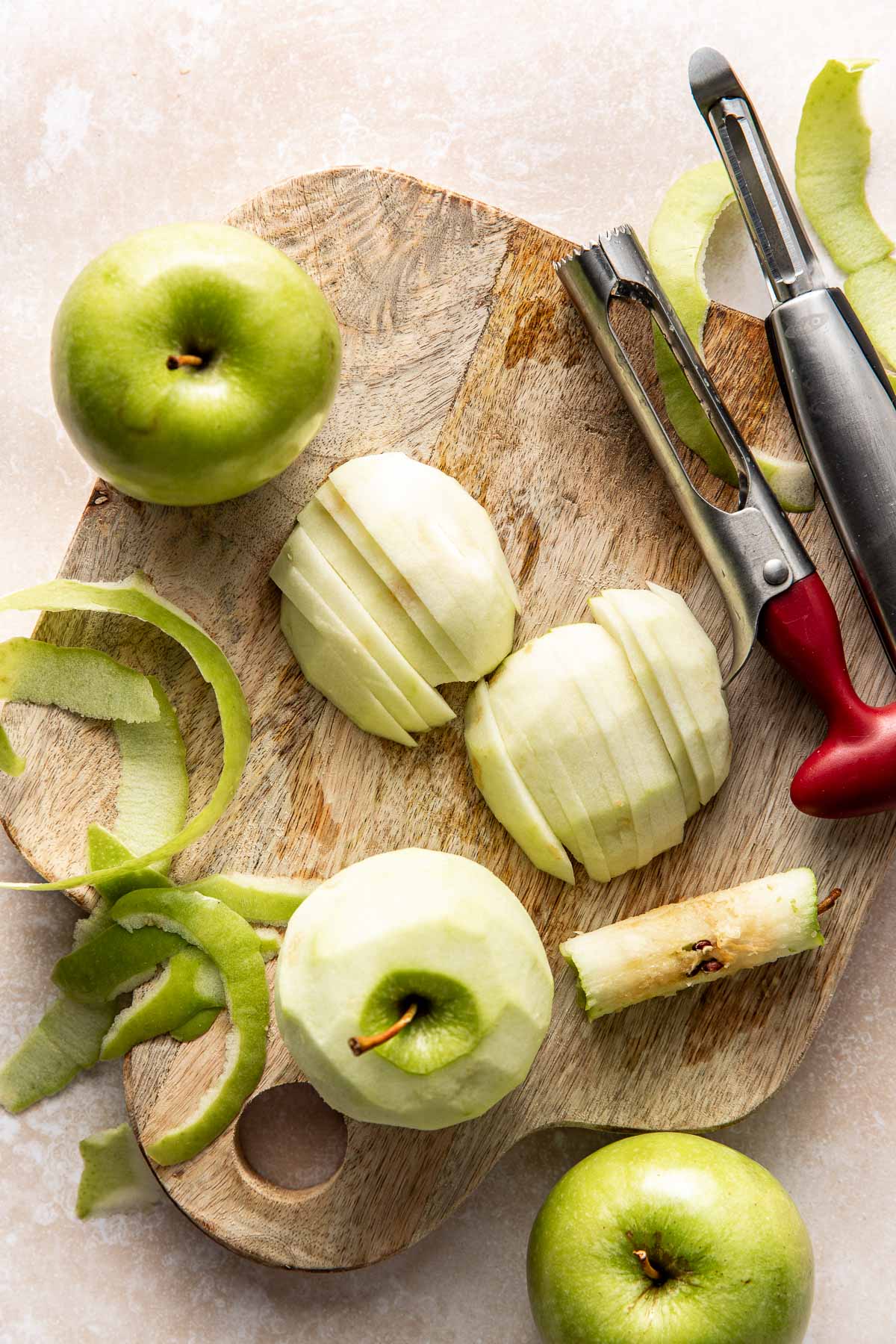 A wooden cutting board with peeled and sliced green apples, apple peels, a corer, and a peeler resting on top, surrounded by whole green apples on a light surface.