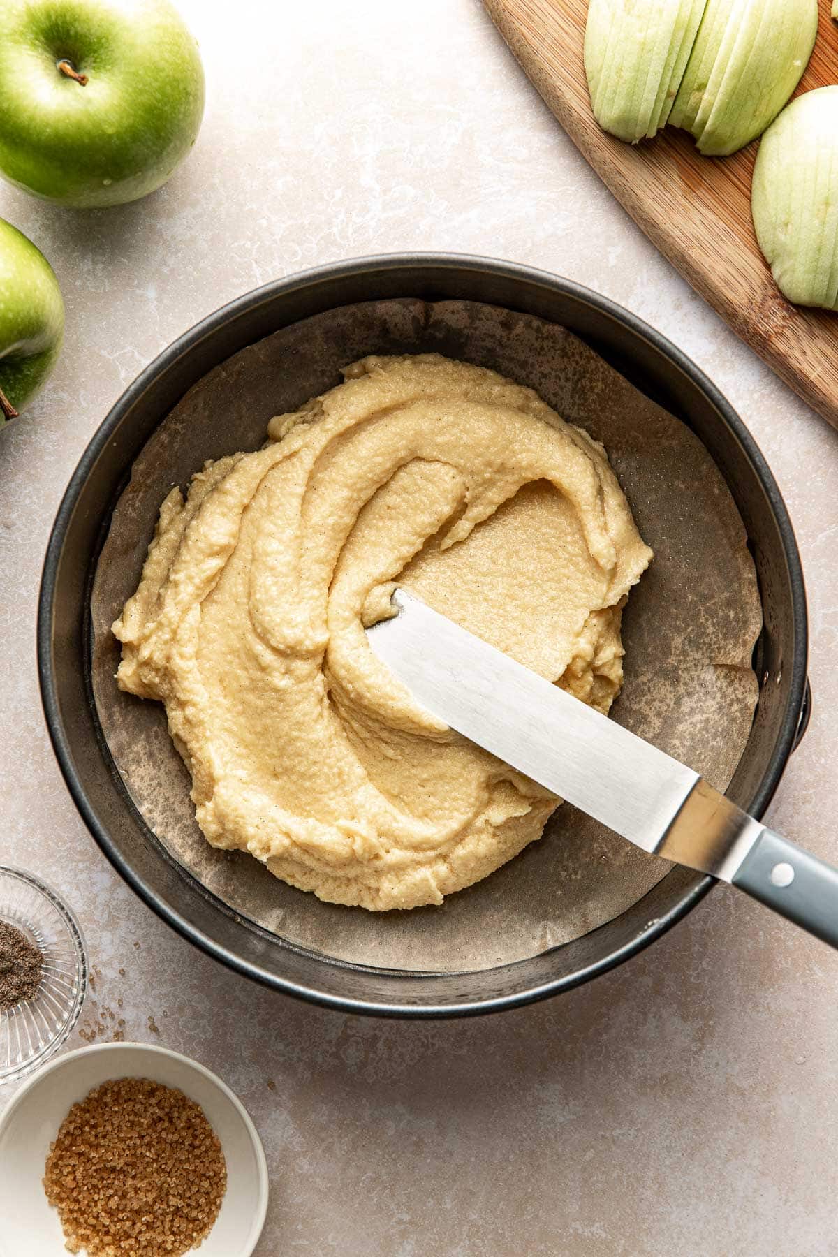 A round cake pan with a layer of batter being spread evenly with a spatula; nearby are green apples, a bowl of brown sugar, a bowl of spices, and sliced apples on a wooden board.