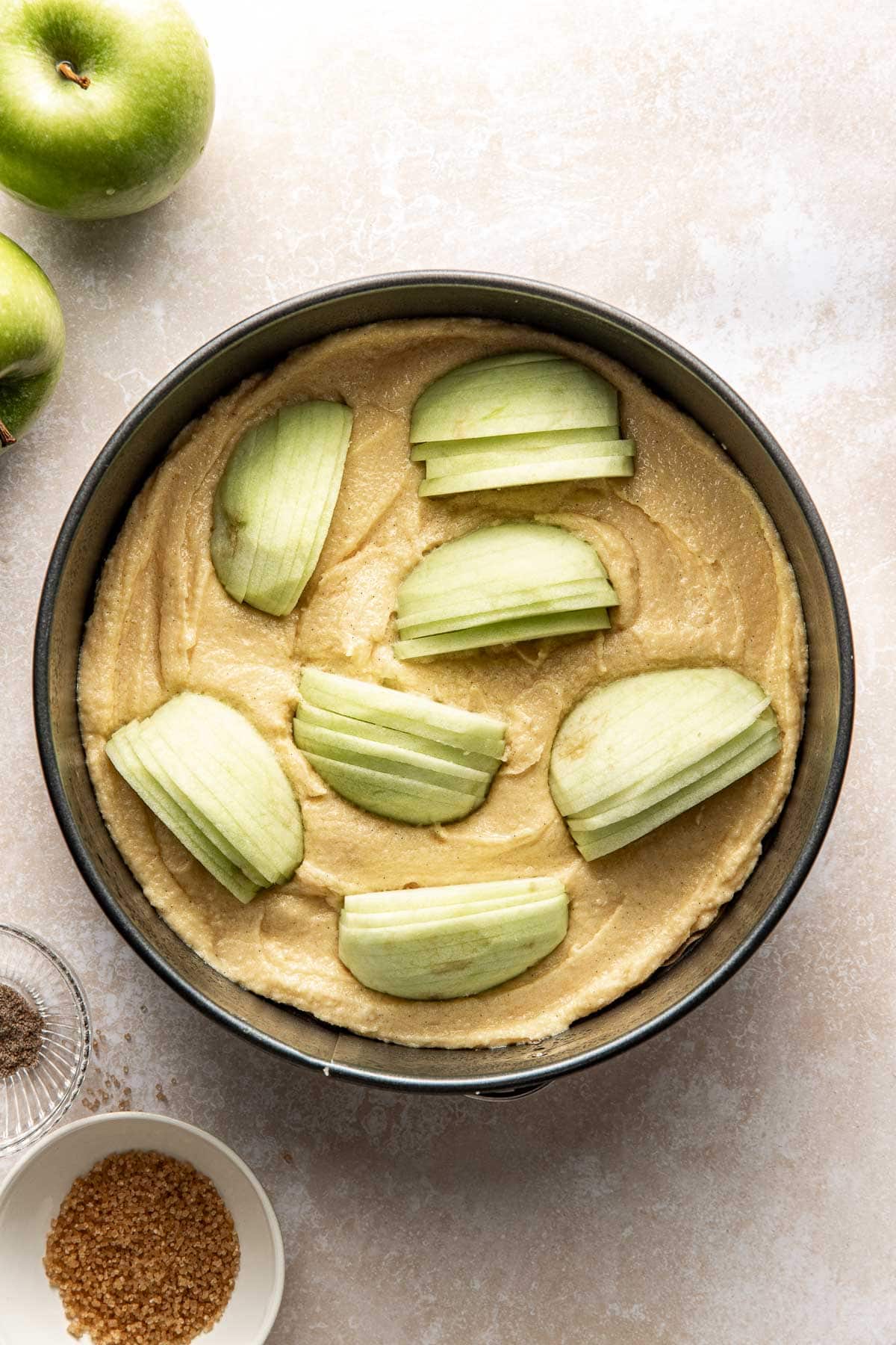 A round cake pan filled with raw batter, topped with sliced green apples arranged in a circular pattern. Nearby are whole green apples, a small bowl of brown sugar, and a small bowl of ground spices.