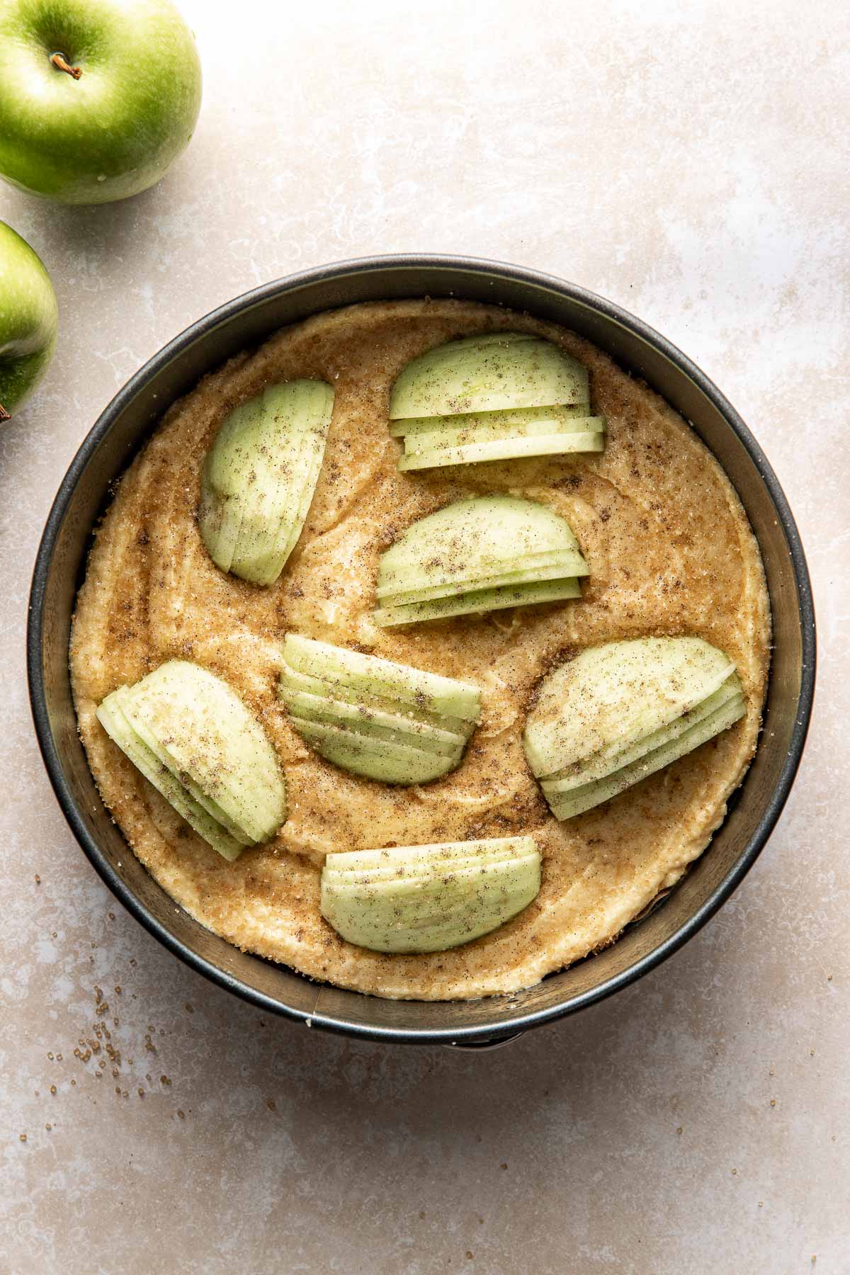 A round baking pan filled with unbaked batter, topped with sliced green apples and sprinkled with spices. Two whole green apples are on the light-colored countertop beside the pan.