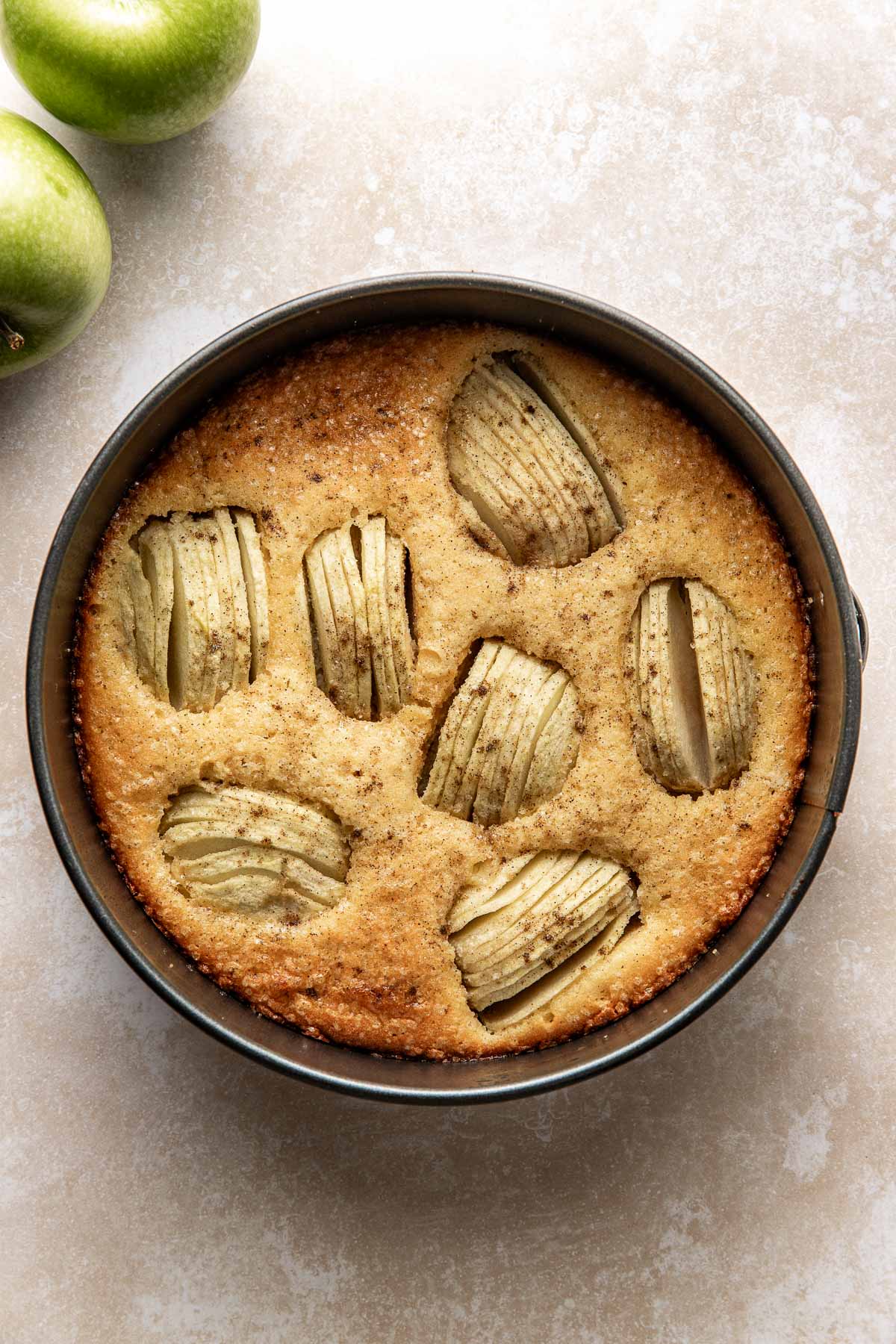 A round apple cake with sliced apples baked on top, sitting in a springform pan on a light-colored surface. Two green apples are visible in the upper left corner.