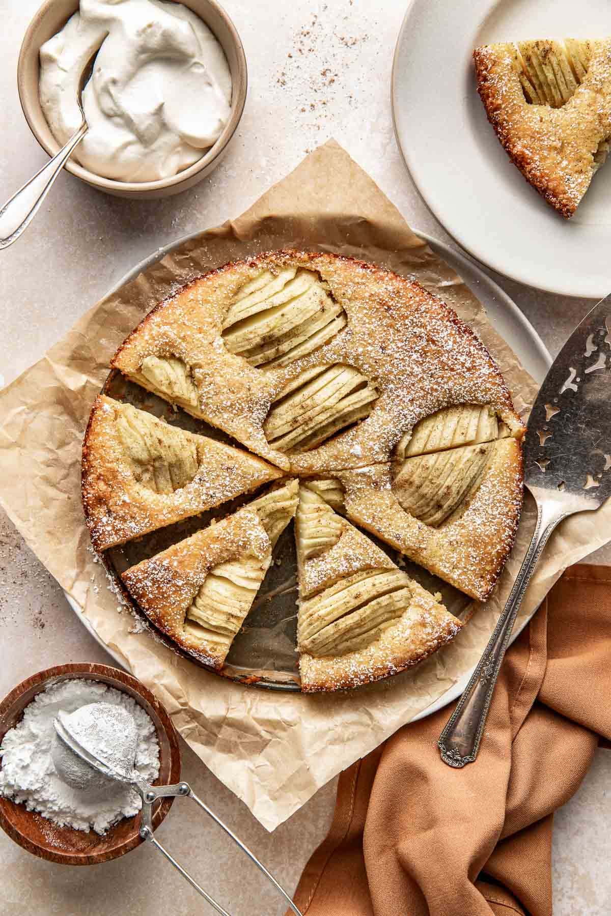 A round apple cake dusted with powdered sugar sits on parchment paper, partially sliced with a serving spatula nearby. A bowl of whipped cream, a small bowl of powdered sugar, and a plate with a cake slice are also shown.