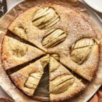 A round pear cake topped with sliced pears, cut into wedges and dusted with powdered sugar, sits on parchment paper with a knife nearby. Bowls of cream and sugar are visible at the edges.