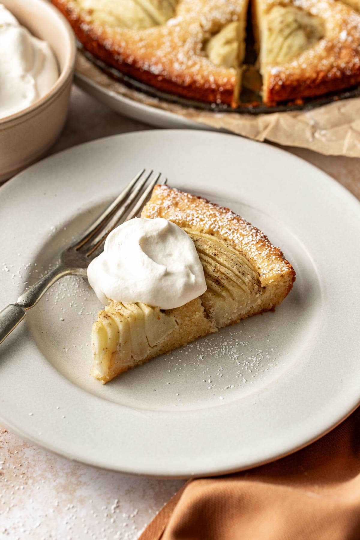 A slice of apple cake topped with a dollop of whipped cream sits on a white plate with a fork, next to a whole cake and a bowl of whipped cream in the background.