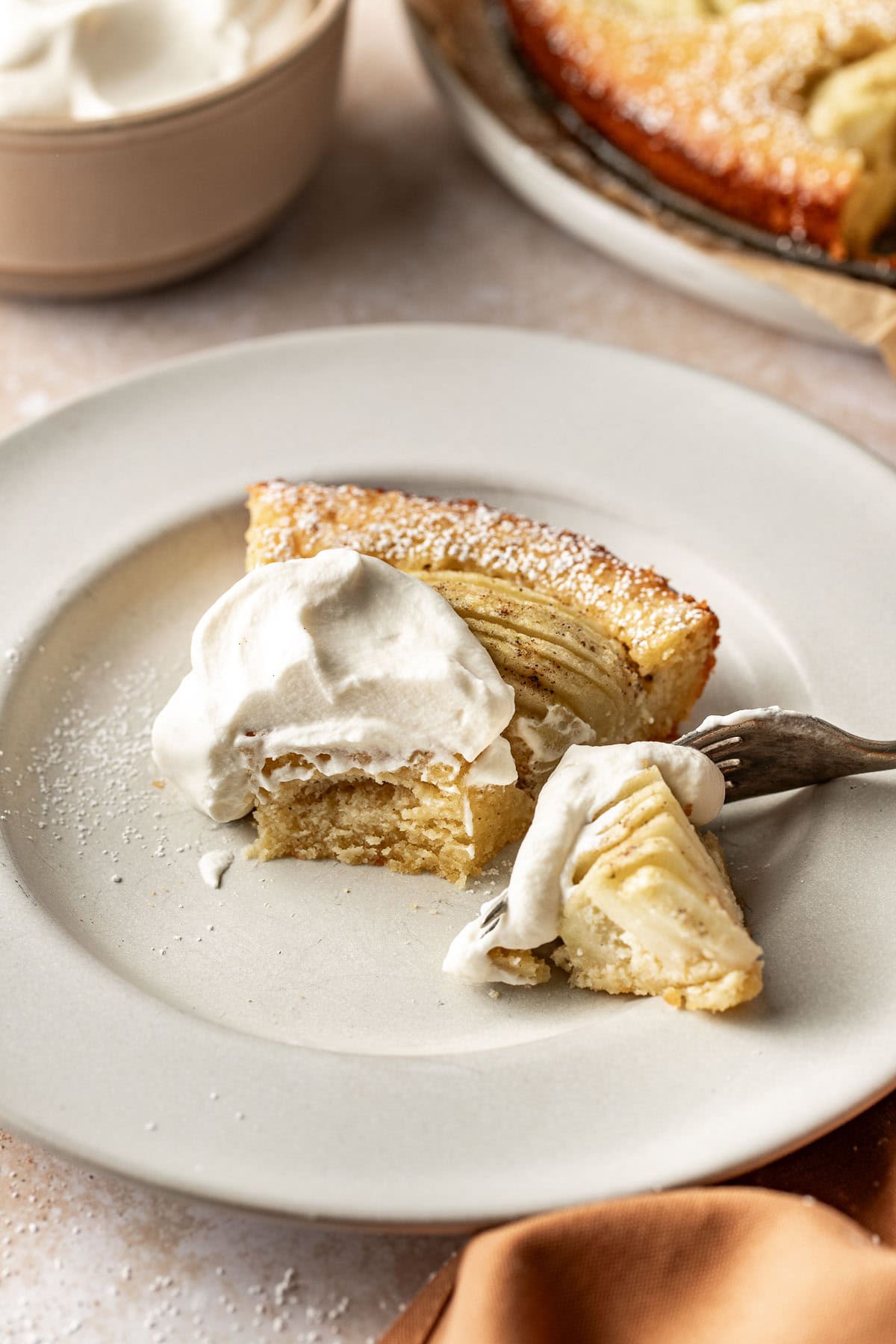 A slice of apple cake topped with whipped cream sits on a white plate. A fork holds a bite of the cake, and more cake is visible in the background along with a bowl of whipped cream.