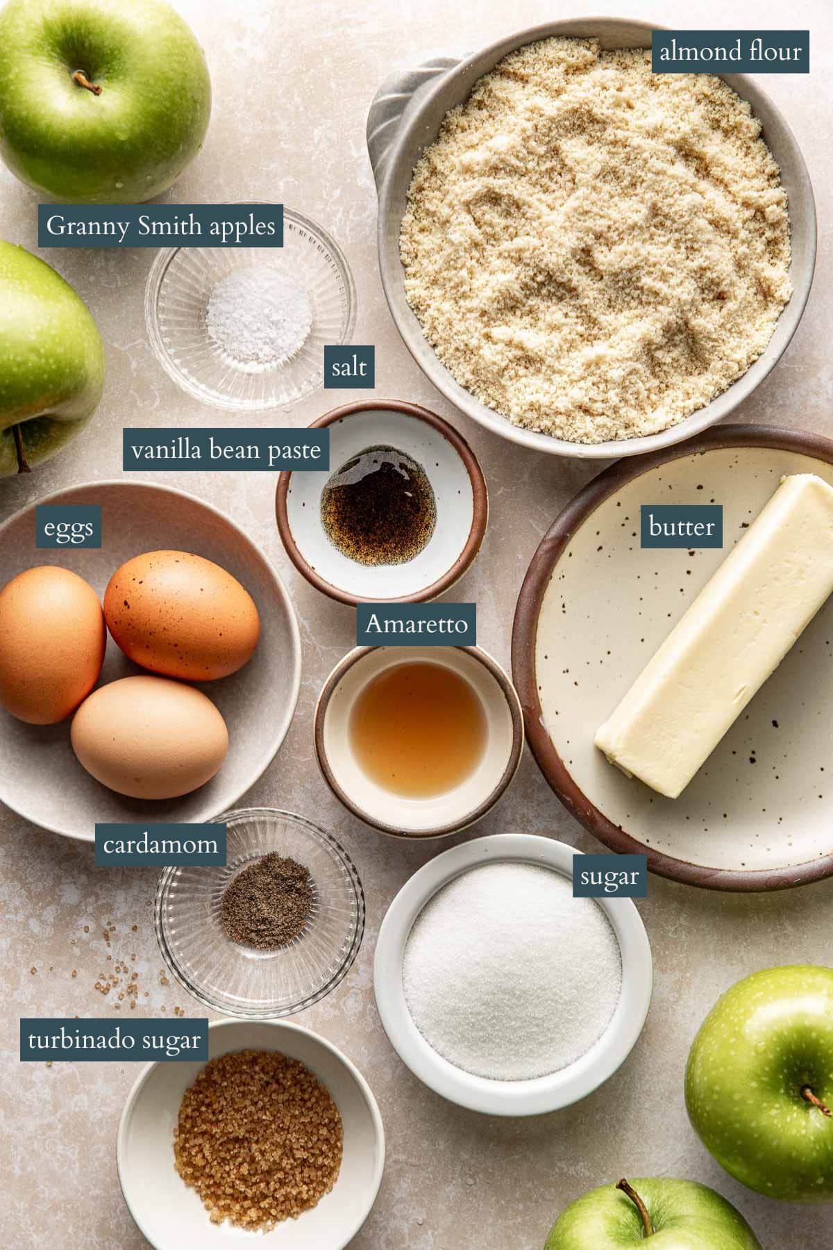 An overhead view of baking ingredients including almond flour, Granny Smith apples, eggs, butter, sugar, turbinado sugar, cardamom, salt, vanilla bean paste, and Amaretto, all labeled on a light surface.