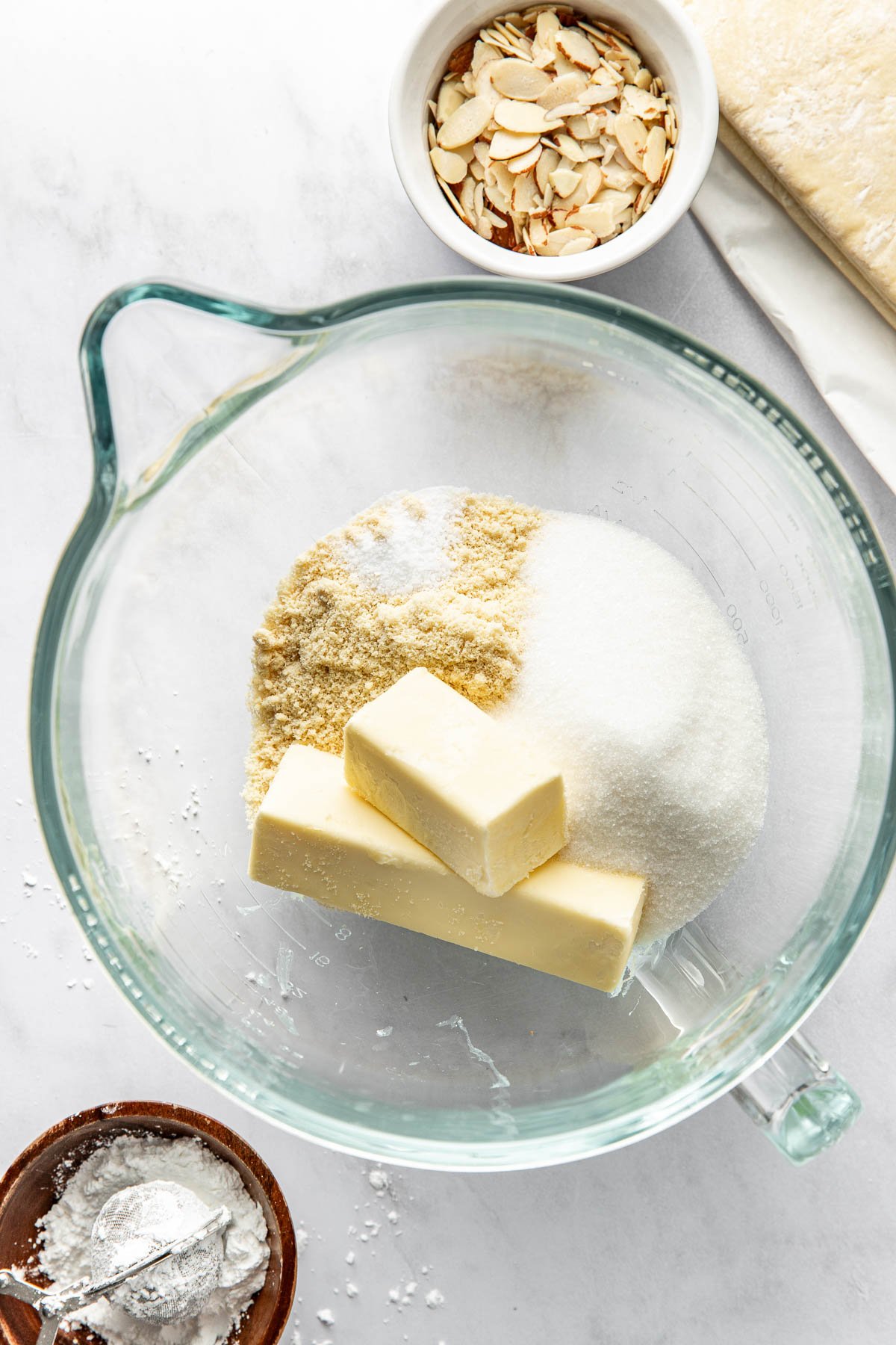 A glass mixing bowl with sugar, almond flour, and two sticks of butter. Nearby are a small bowl of sliced almonds, a small bowl of flour with a spoon, and a piece of dough on a white surface.
