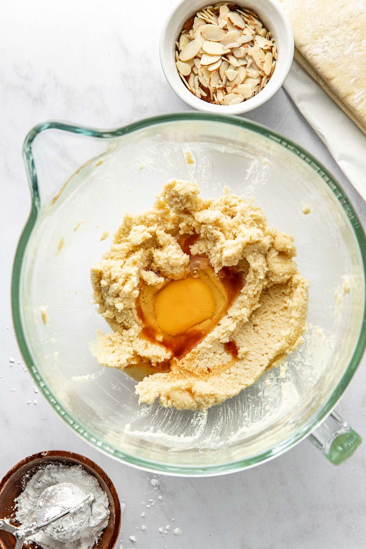 A glass mixing bowl with creamed butter and sugar, an egg, and vanilla extract. Nearby are a bowl of sliced almonds, a small bowl with a spoon and flour, and a sheet of dough on a cloth.