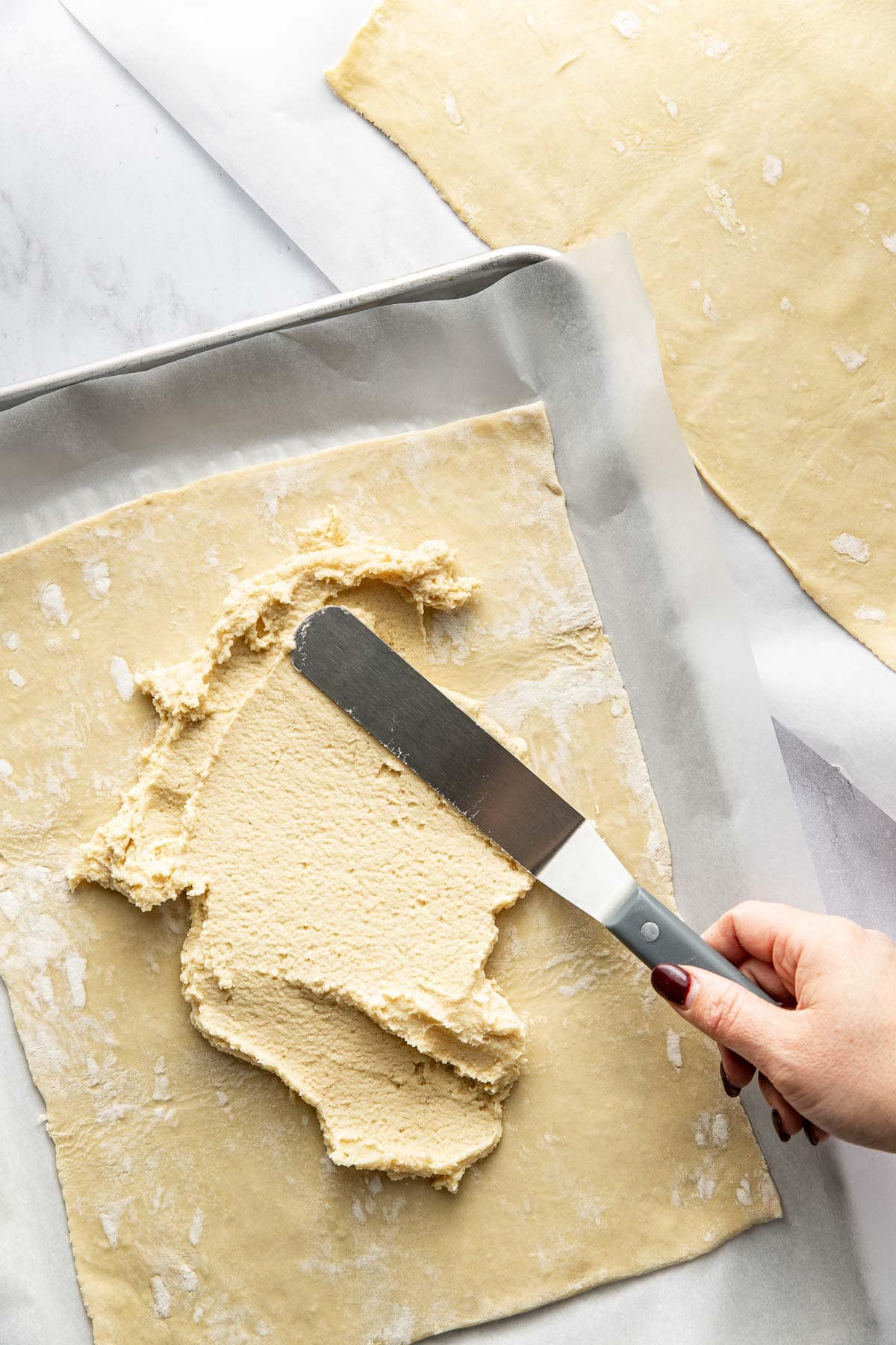 A hand spreads a creamy filling onto a large sheet of raw pastry dough with an offset spatula, on a parchment-lined baking tray. Another sheet of dough is visible in the background.