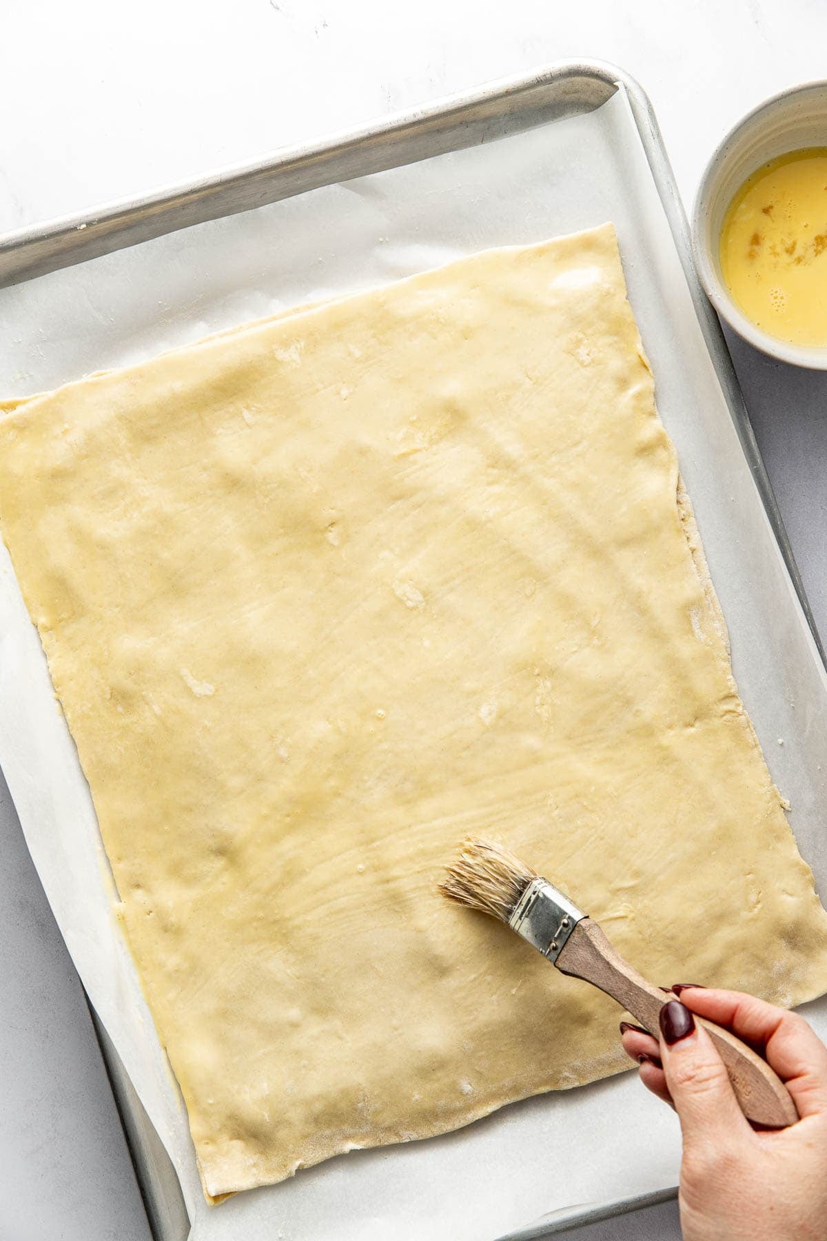 A hand uses a pastry brush to apply an egg wash to a large sheet of dough on a baking tray lined with parchment paper. A bowl of egg wash sits nearby.