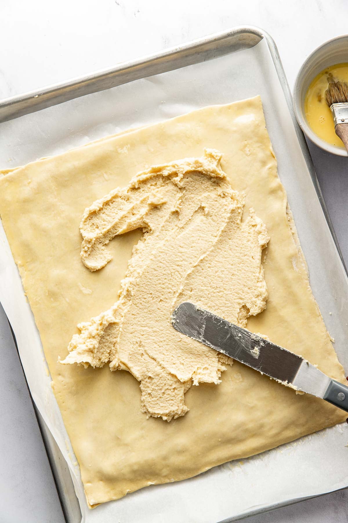 A baking sheet lined with parchment paper holds a sheet of pastry dough. An offset spatula spreads a creamy filling over the dough, with a bowl of egg wash and a pastry brush nearby.