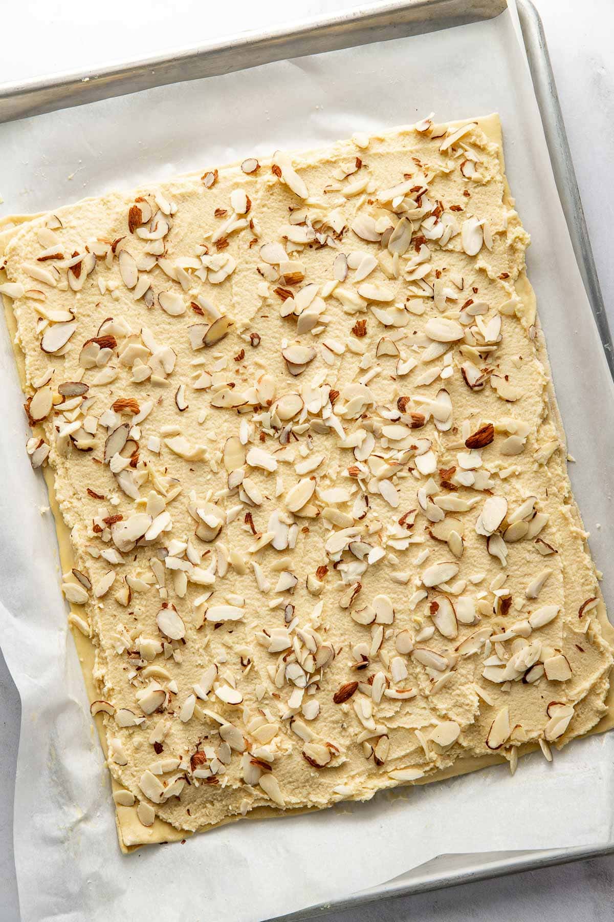 A rectangular sheet of dough covered with a creamy filling and topped with sliced almonds, placed on parchment paper on a baking tray.