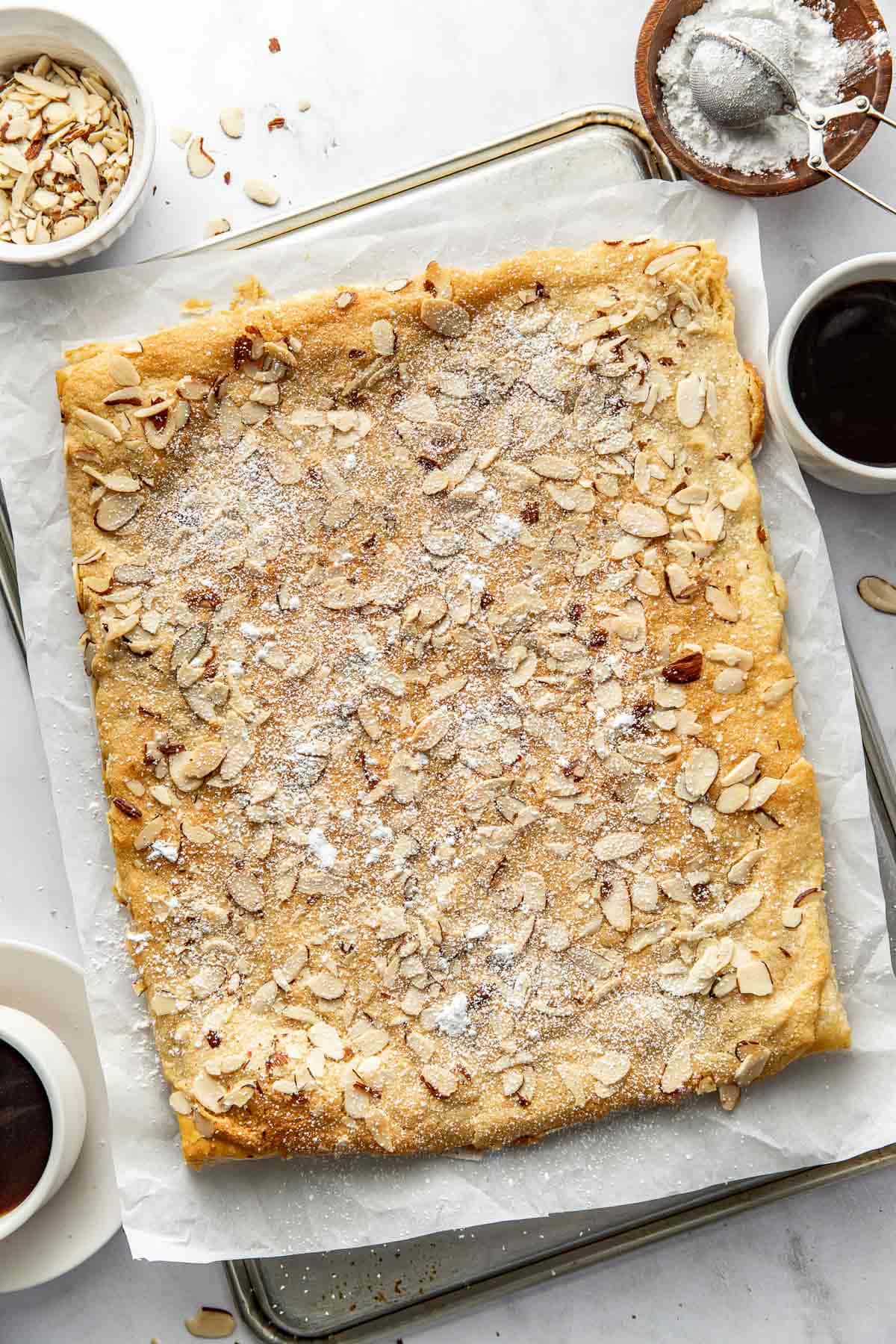 A rectangular almond pastry topped with sliced almonds and powdered sugar sits on parchment paper. Surrounding it are cups of coffee, a small bowl of almonds, and a bowl of powdered sugar.