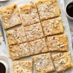 A tray of sliced almond pastry squares topped with powdered sugar and slivered almonds, surrounded by coffee cups, a bowl of powdered sugar, and a small dish of almonds on a light background.