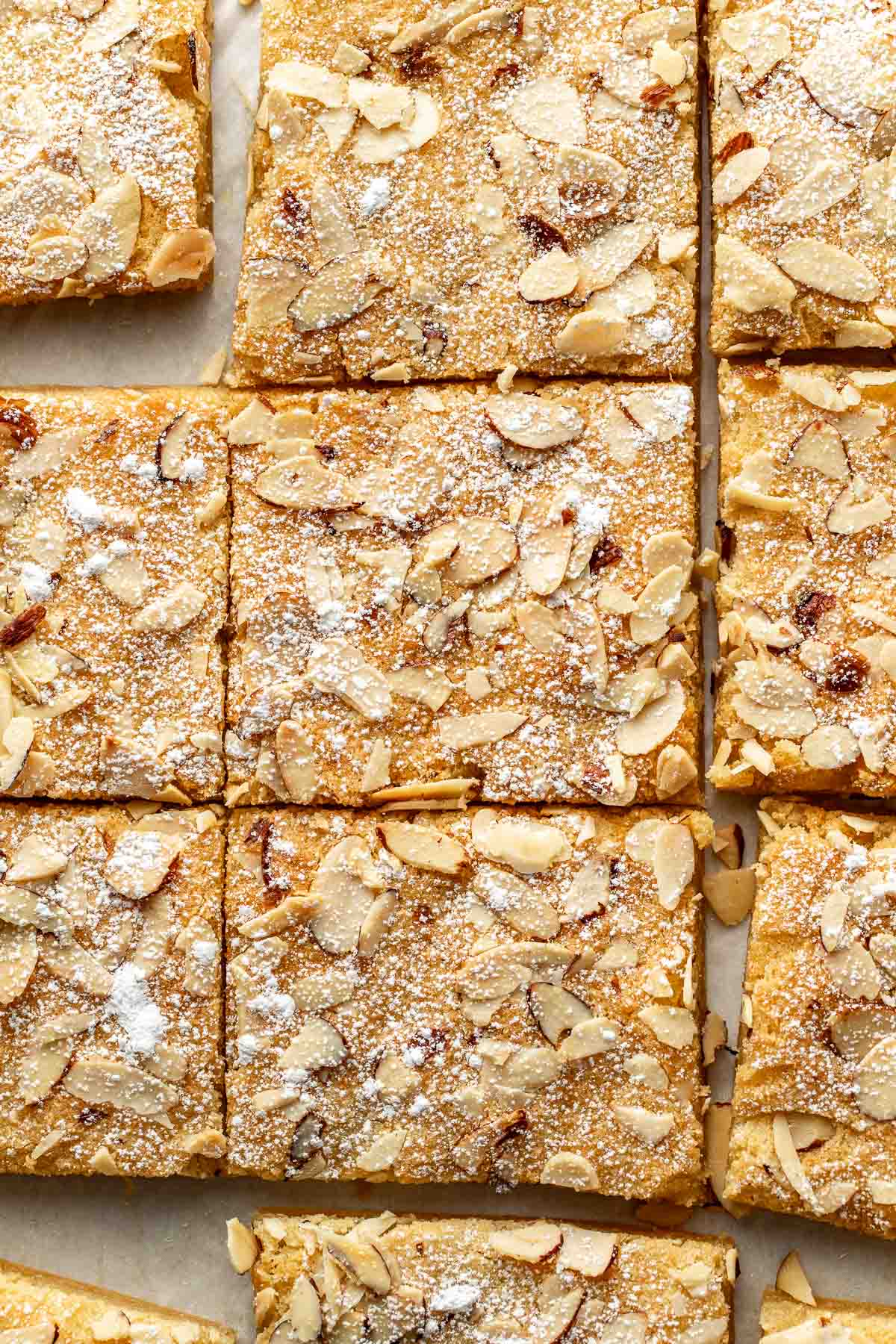 A close-up of sliced almond bars topped with powdered sugar and slivered almonds, arranged in neat squares on a light surface.