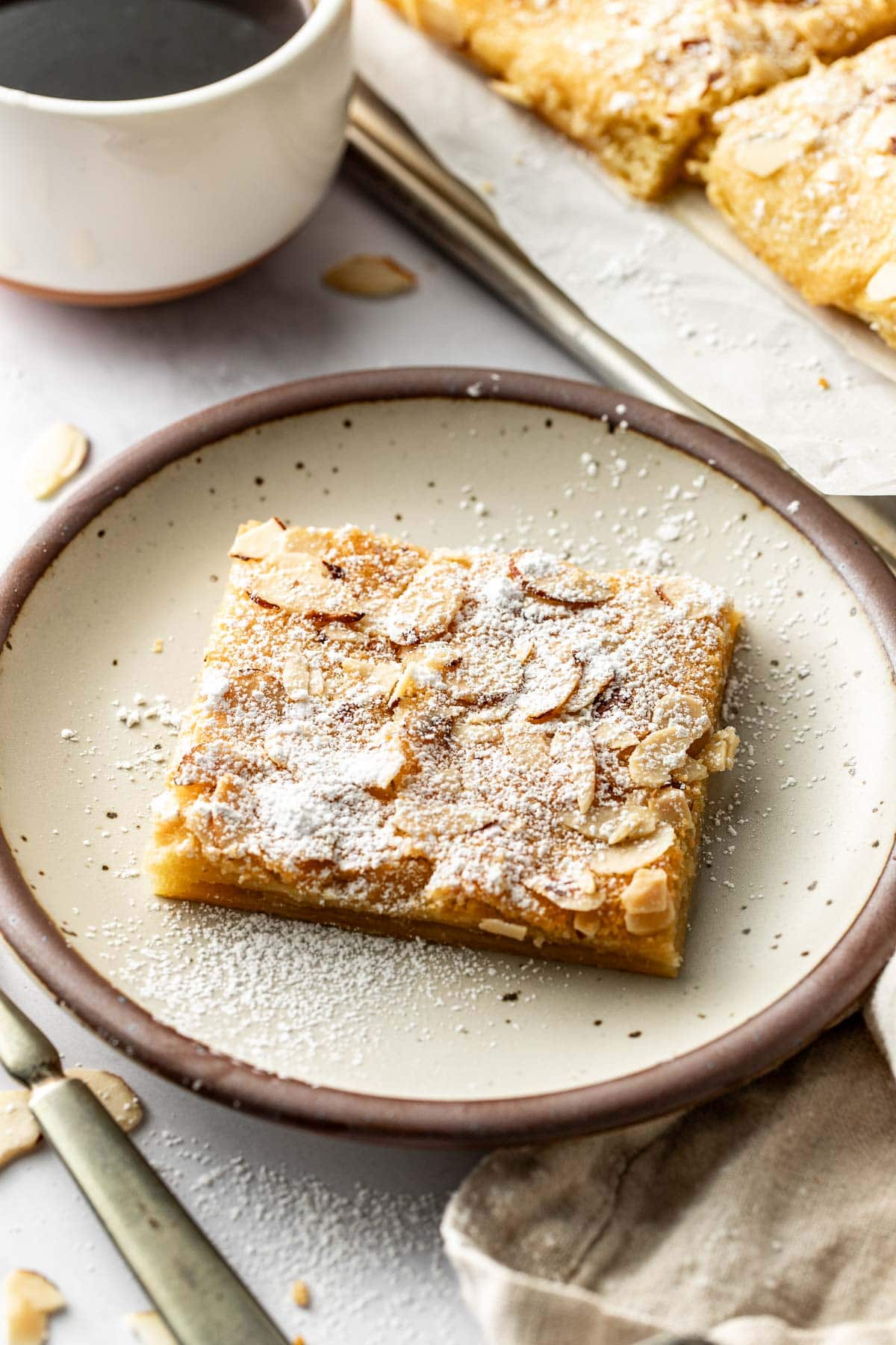 A square almond pastry dusted with powdered sugar sits on a plate, with a knife, napkin, and a cup of coffee nearby. More pastries are visible in the background.