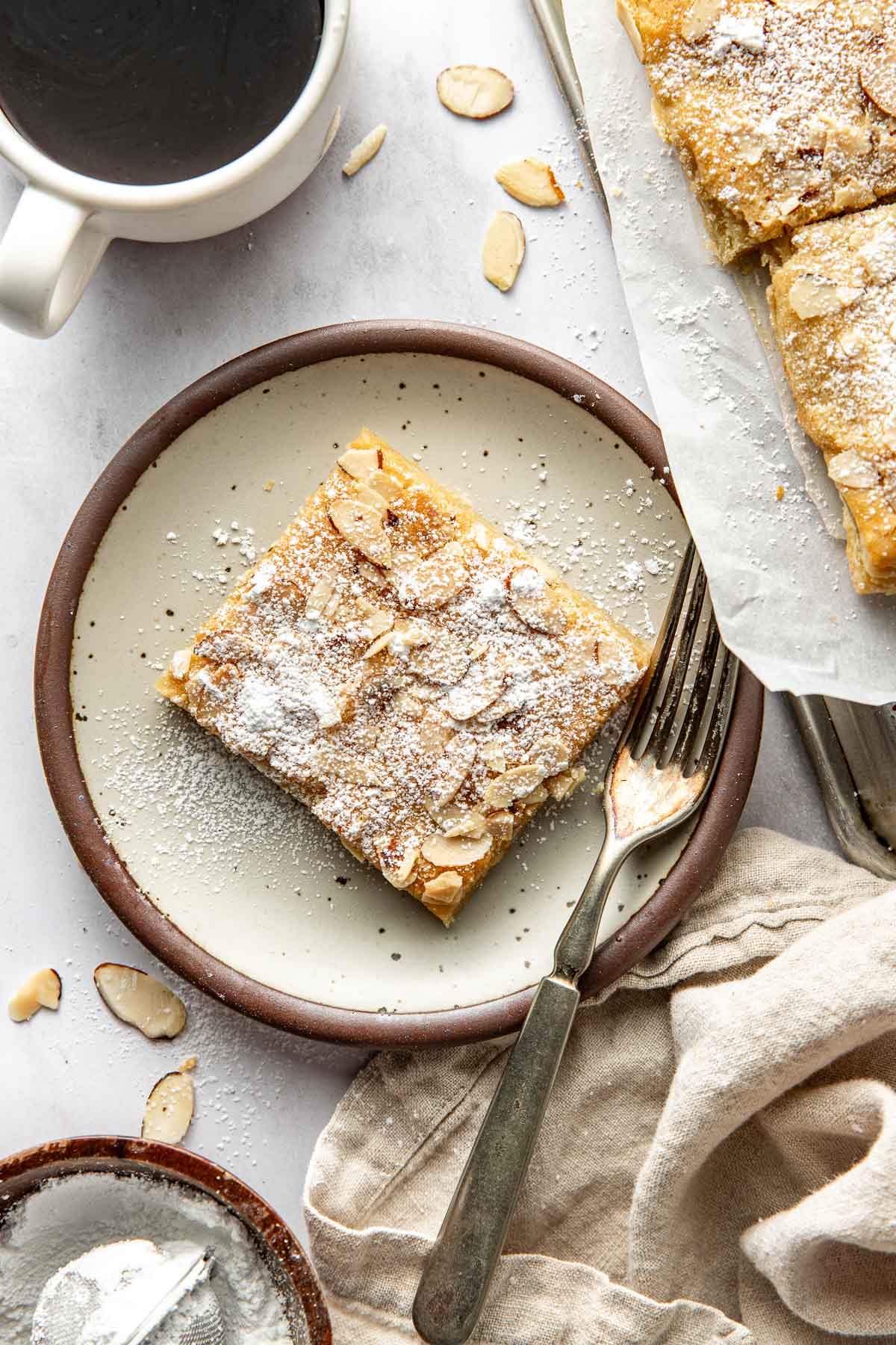 A square slice of almond pastry dusted with powdered sugar sits on a plate with a fork. Scattered almond slices, a cup of coffee, and a napkin are nearby on a light surface.