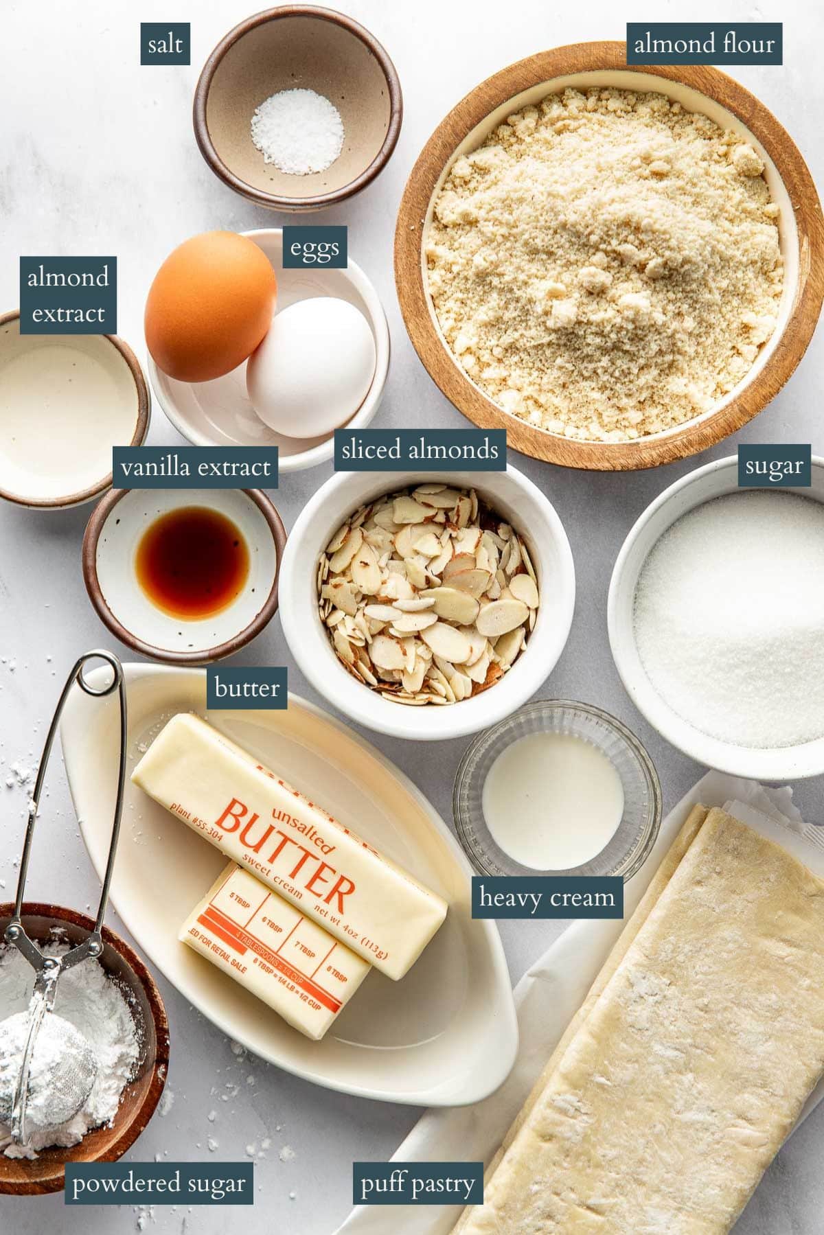 A top-down view of baking ingredients, each labeled: salt, eggs, almond extract, vanilla extract, butter, powdered sugar, almond flour, sliced almonds, heavy cream, sugar, and puff pastry.