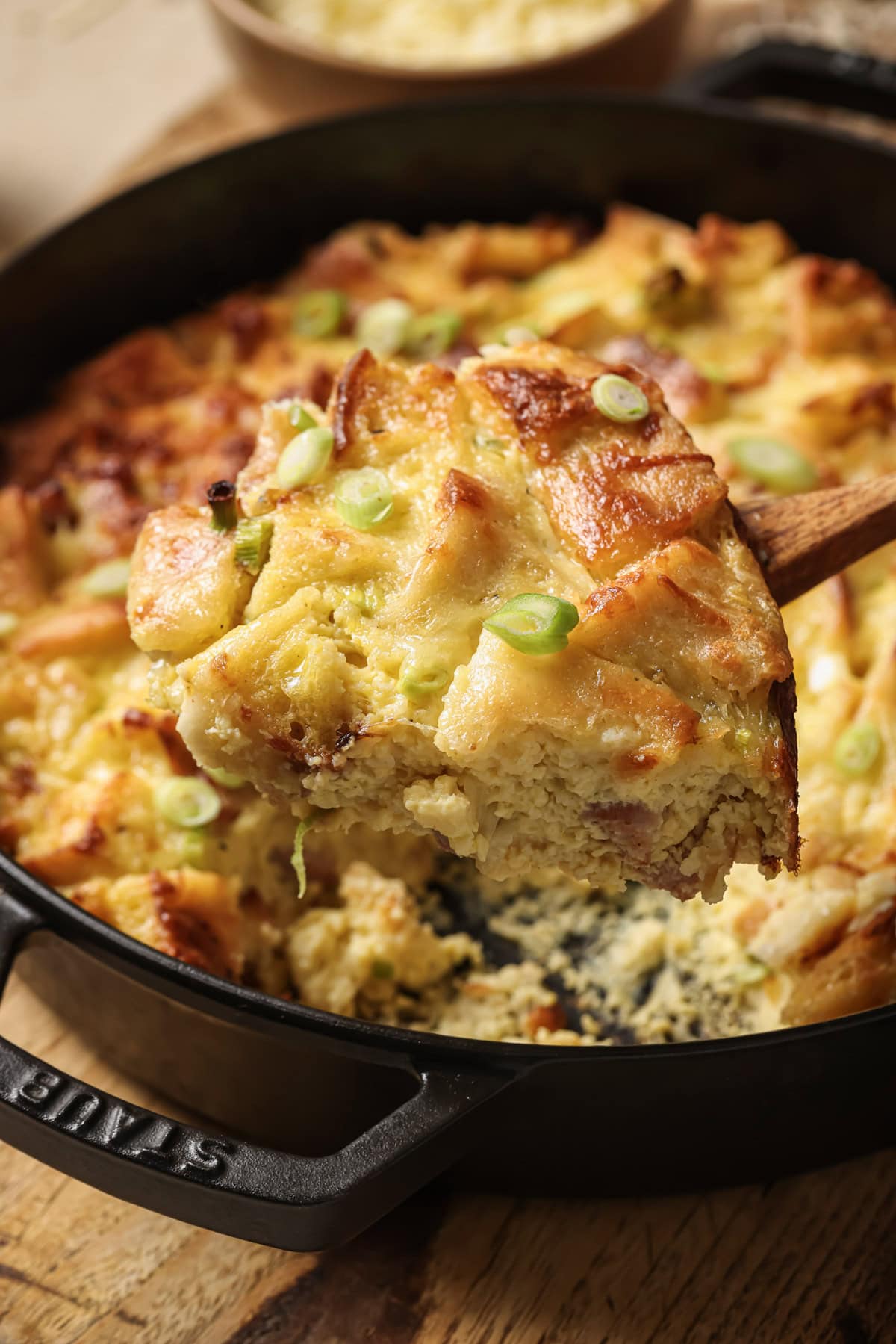 A close-up of a casserole dish with a golden, cheesy bread pudding topped with sliced green onions. A wooden spatula lifts a serving, showing a soft, creamy interior and crispy browned top.