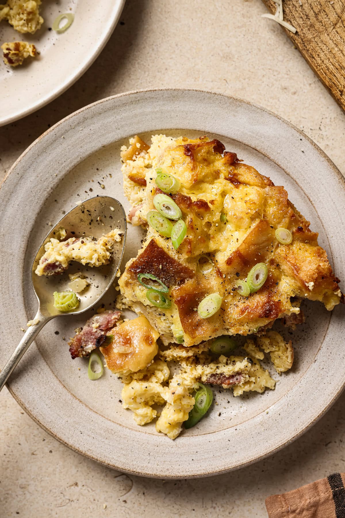A slice of savory bread pudding topped with sliced green onions sits on a beige plate, with a spoon beside it. The dish has a golden, crispy top and a creamy interior with visible pieces of sausage.