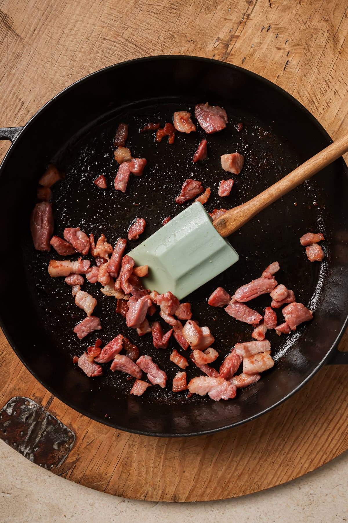 A green spatula stirs small pieces of raw bacon in a black skillet on a wooden surface.