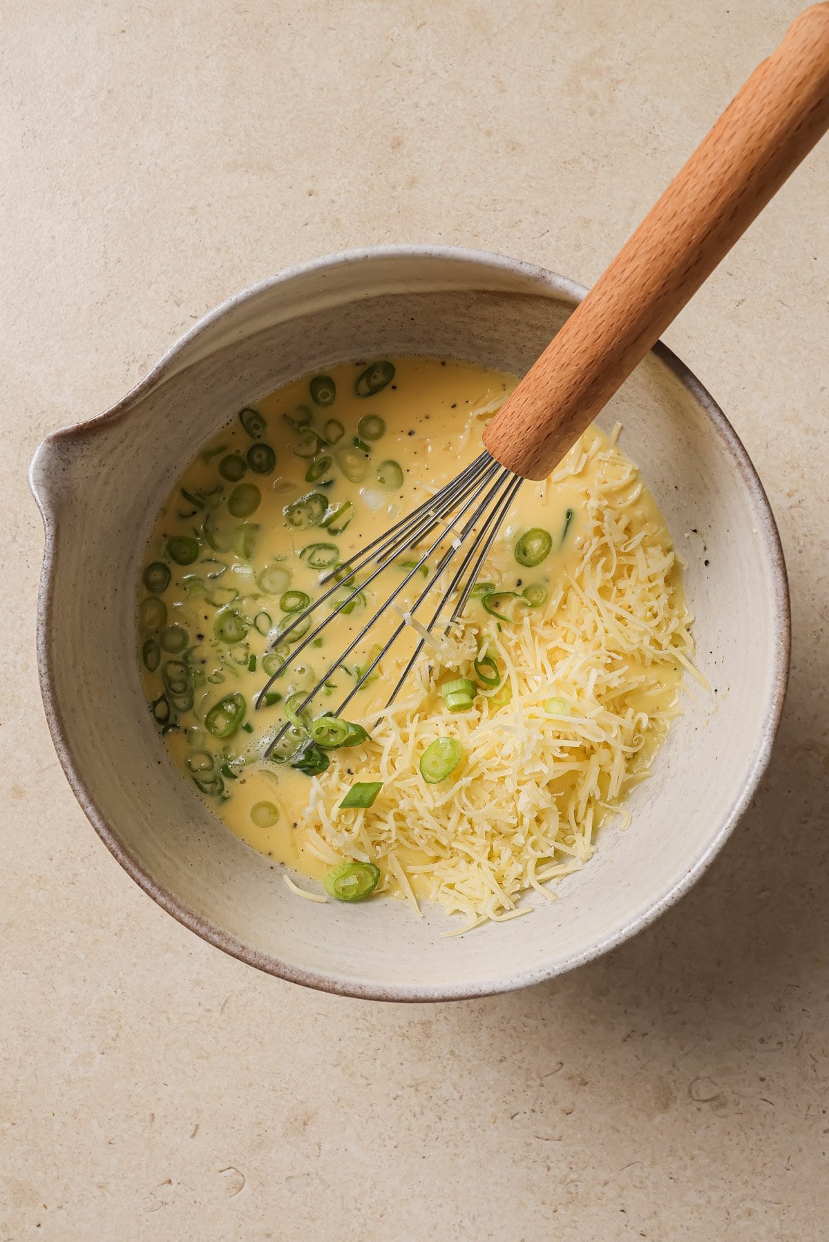 A mixing bowl containing beaten eggs, shredded cheese, and sliced green onions, with a metal whisk with a wooden handle resting inside. The bowl sits on a light-colored countertop.