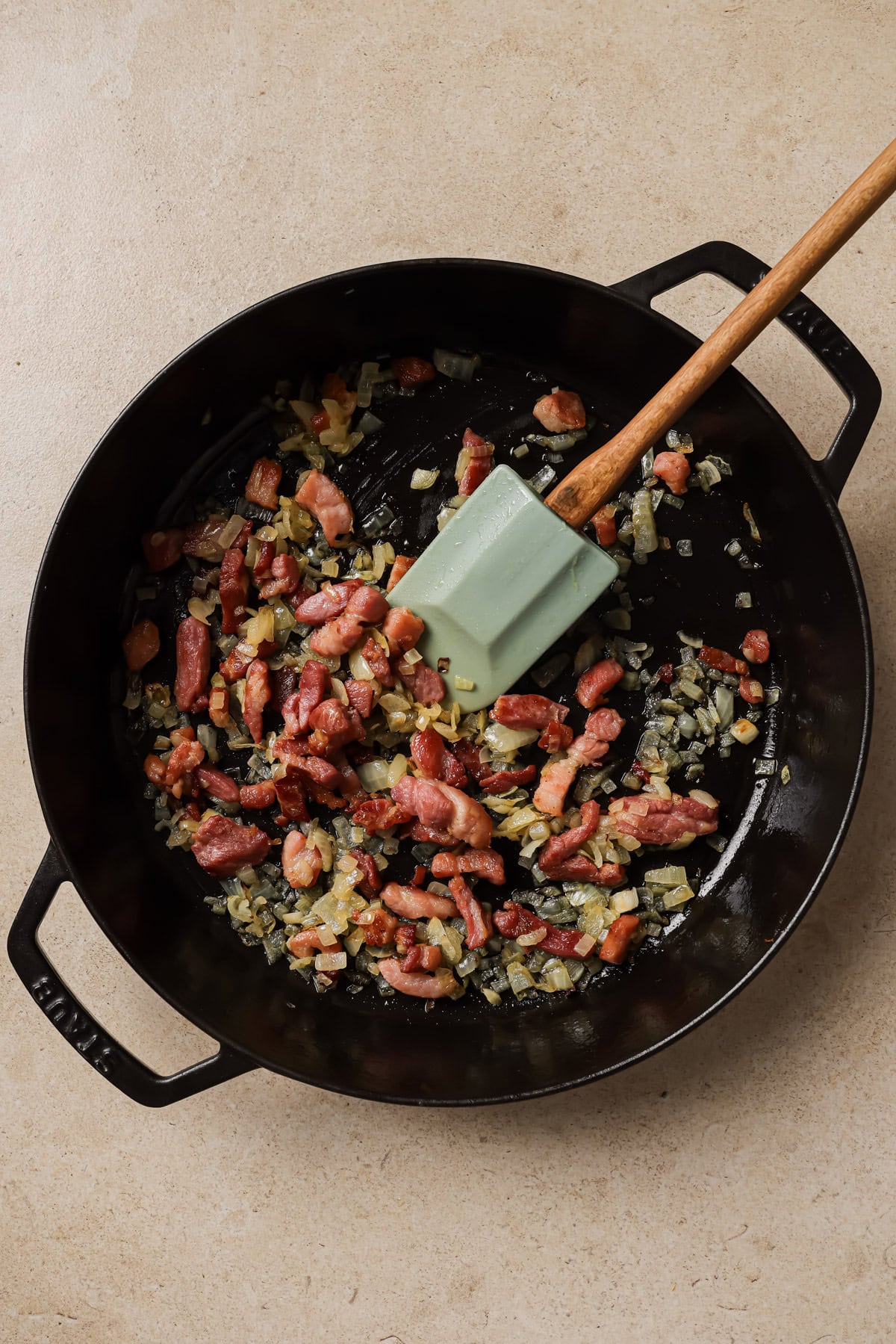 A black skillet with chopped onions and bacon cooking inside, being stirred with a green spatula on a beige countertop.