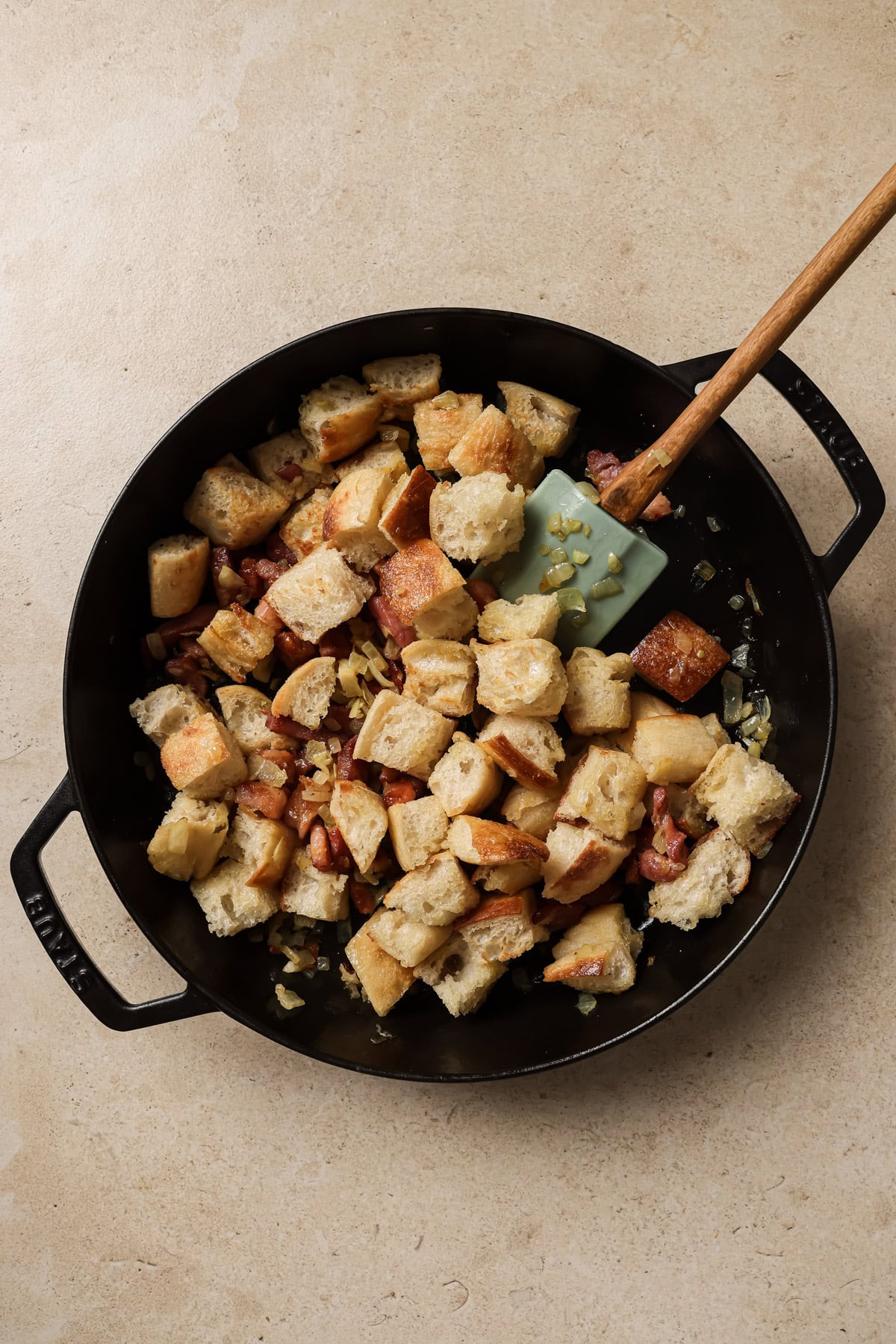 A black skillet filled with cubed bread, sautéed onions, and bacon pieces, with a light blue spatula stirring the mixture on a light beige surface.