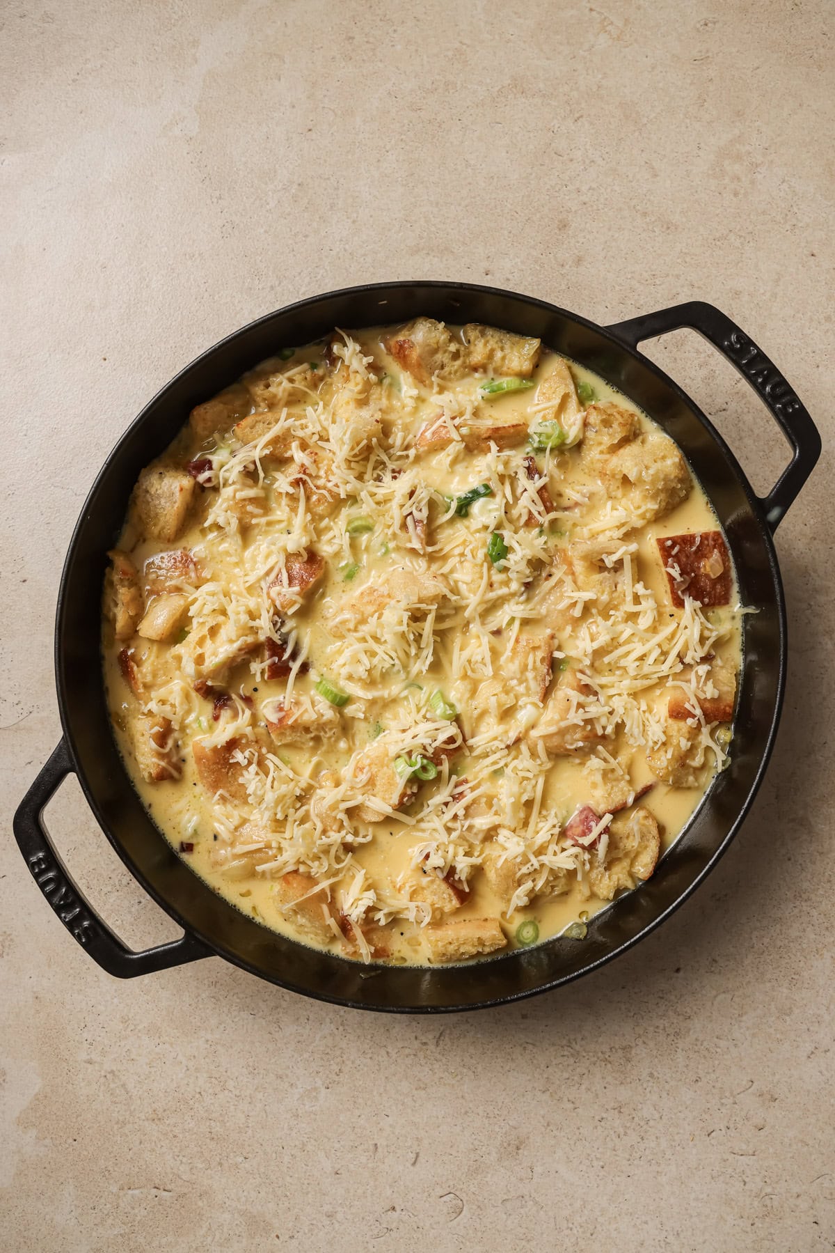 A round black pan filled with a creamy, cheesy casserole featuring chunks of bread, melted cheese, and scattered vegetables, set on a light-colored surface.