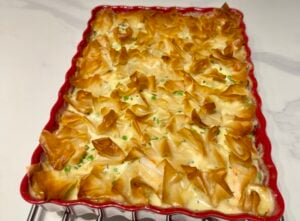 A baked casserole in a red scalloped dish, topped with golden, crispy layers of phyllo pastry. Perfect for January feel-food recipes, its filling reveals hints of green herbs or vegetables, and the dish rests on a white countertop.
