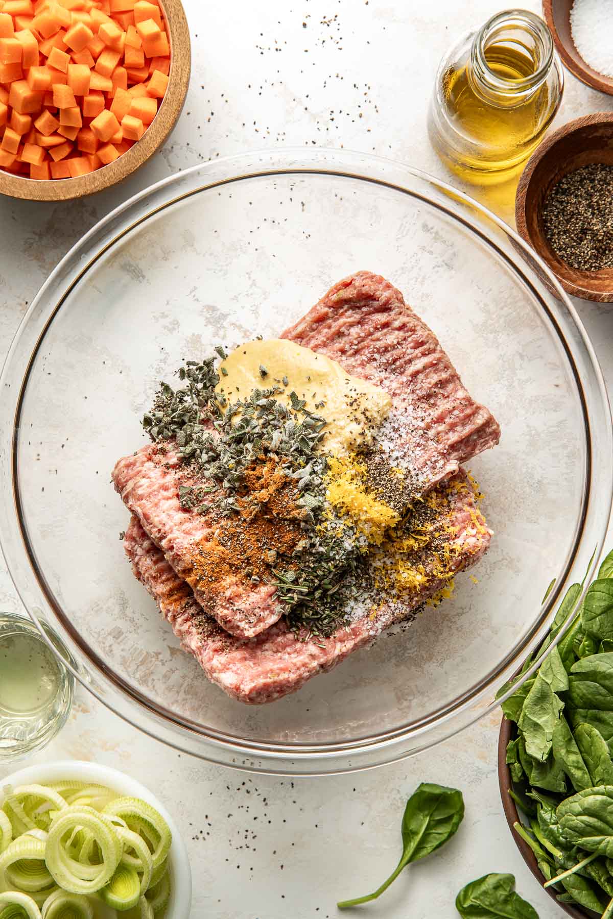 A glass bowl with ground meat, herbs, spices, mustard, and lemon zest on a white surface. Surrounding it are chopped carrots, oil, pepper, fresh spinach, sliced leeks, and a small dish with liquid.