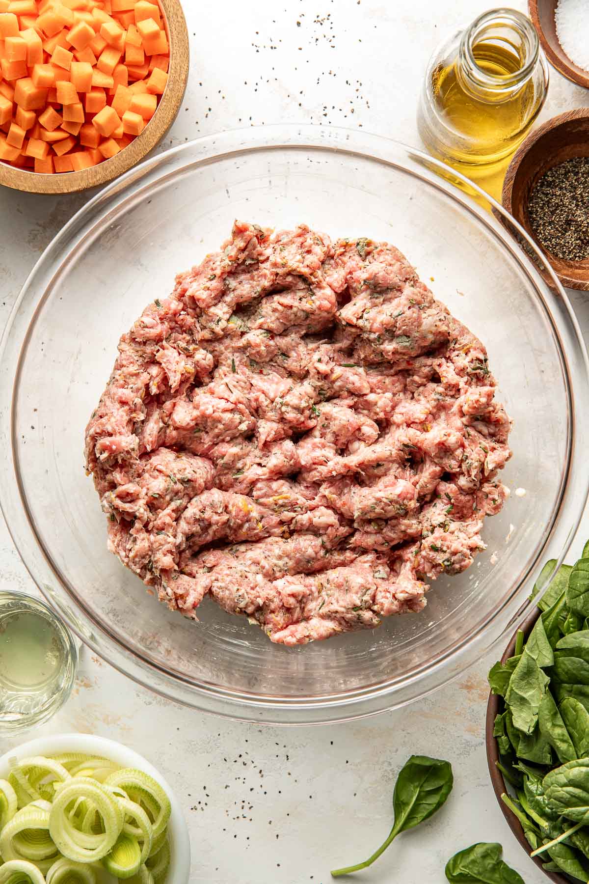 A glass bowl filled with seasoned raw ground meat sits on a white surface, surrounded by bowls of chopped carrots, fresh spinach leaves, sliced leeks, a bottle of olive oil, and a bowl of black pepper.