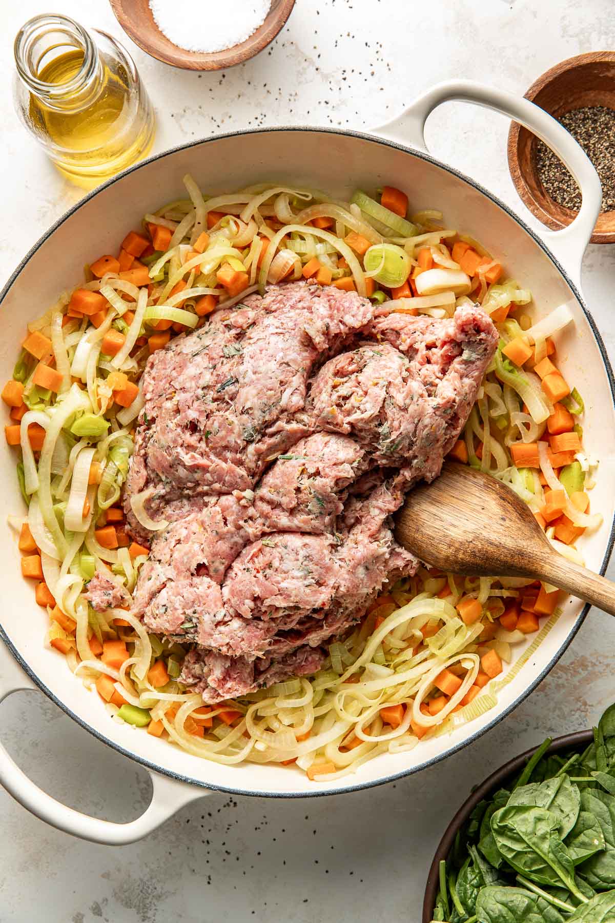 A large pot contains ground turkey on top of sautéed chopped carrots, celery, and onions, with a wooden spoon resting inside. Surrounding the pot are bowls of salt, pepper, olive oil, and fresh spinach leaves.