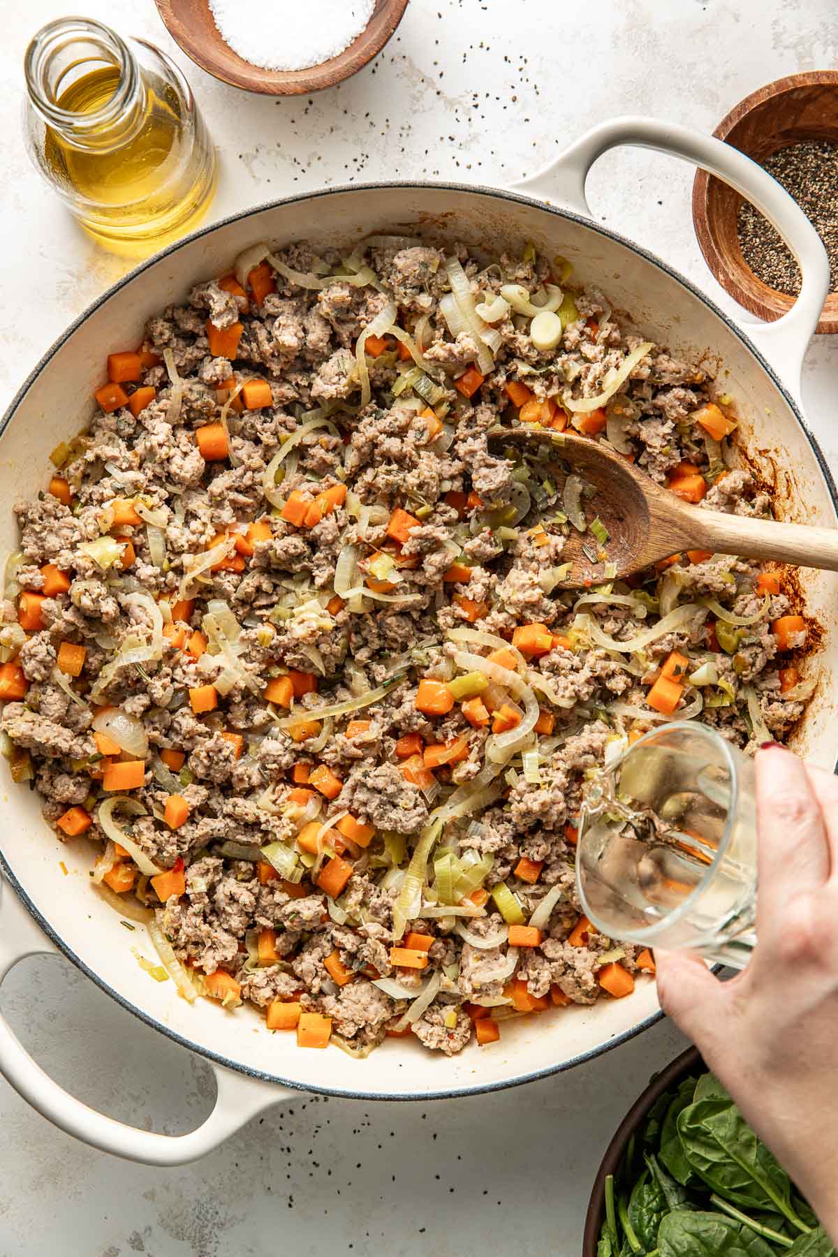 A hand pours liquid from a small glass into a pan filled with cooked ground meat, chopped carrots, and onions. A wooden spoon rests in the pan. Surrounding the pan are bowls of salt, pepper, and fresh spinach, and a bottle of oil.