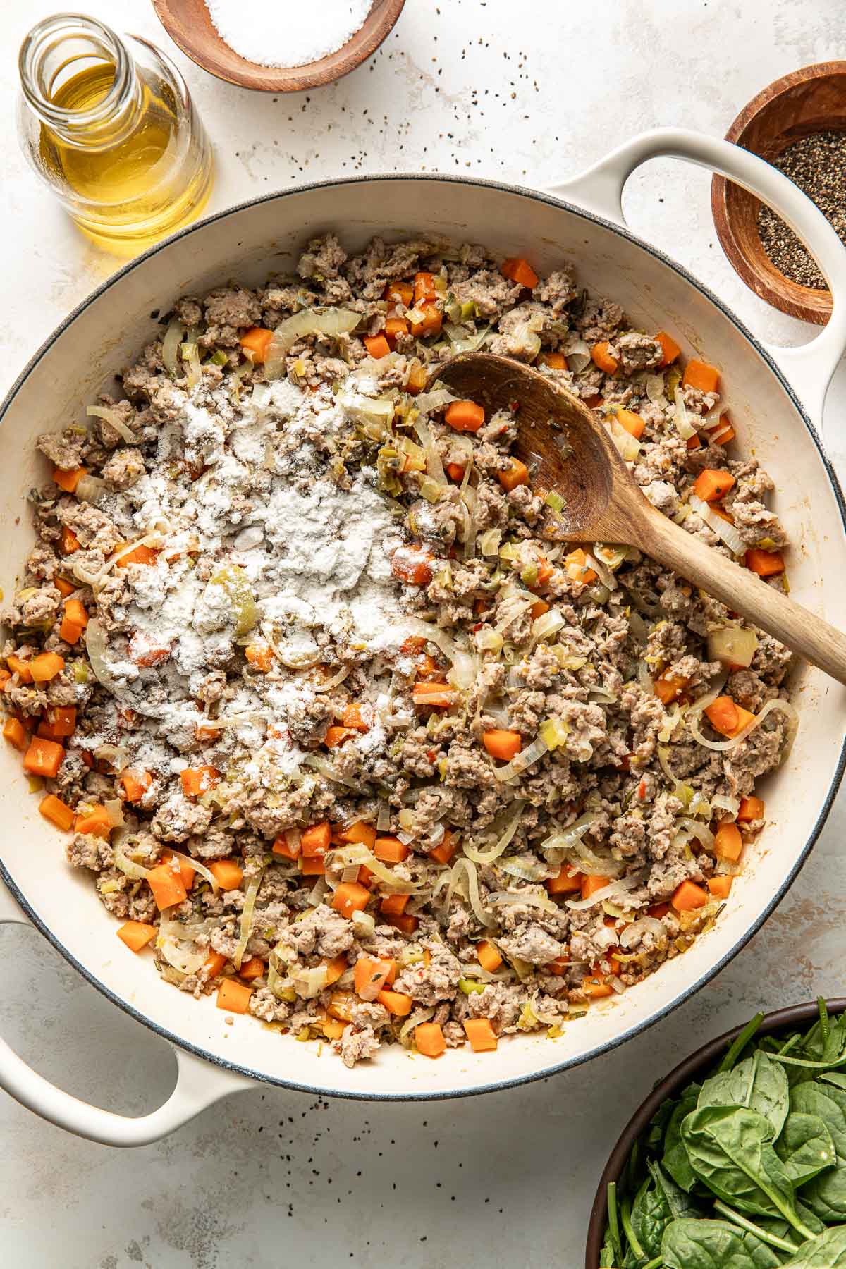 A large white pan filled with ground meat, chopped carrots, onions, and flour being stirred with a wooden spoon. Ingredients like oil, salt, and pepper are nearby on a light countertop.
