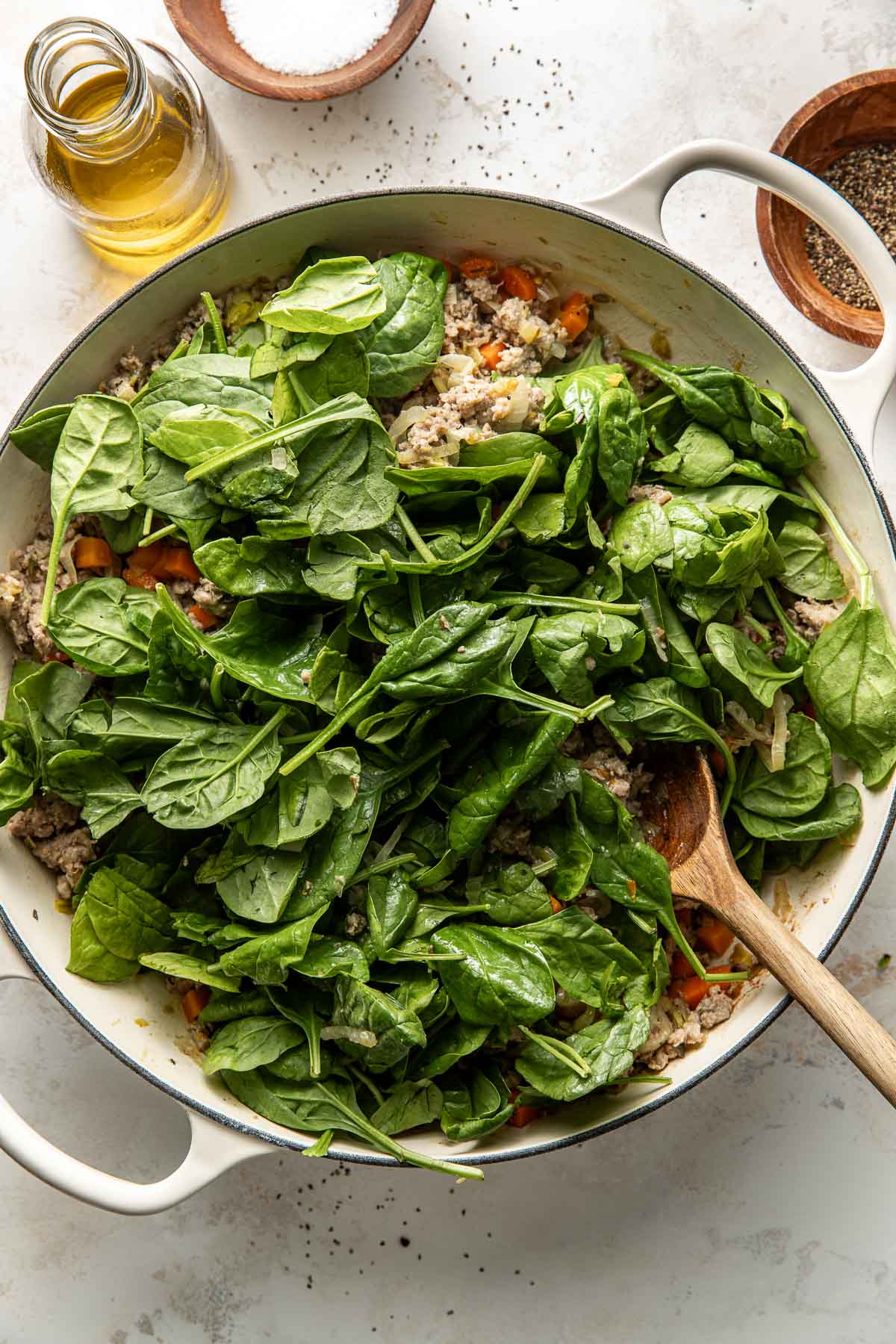 A white pot filled with ground meat, chopped vegetables, and fresh spinach leaves being stirred with a wooden spoon. Nearby are a small bottle of oil, a bowl of salt, and a bowl of pepper on a light surface.