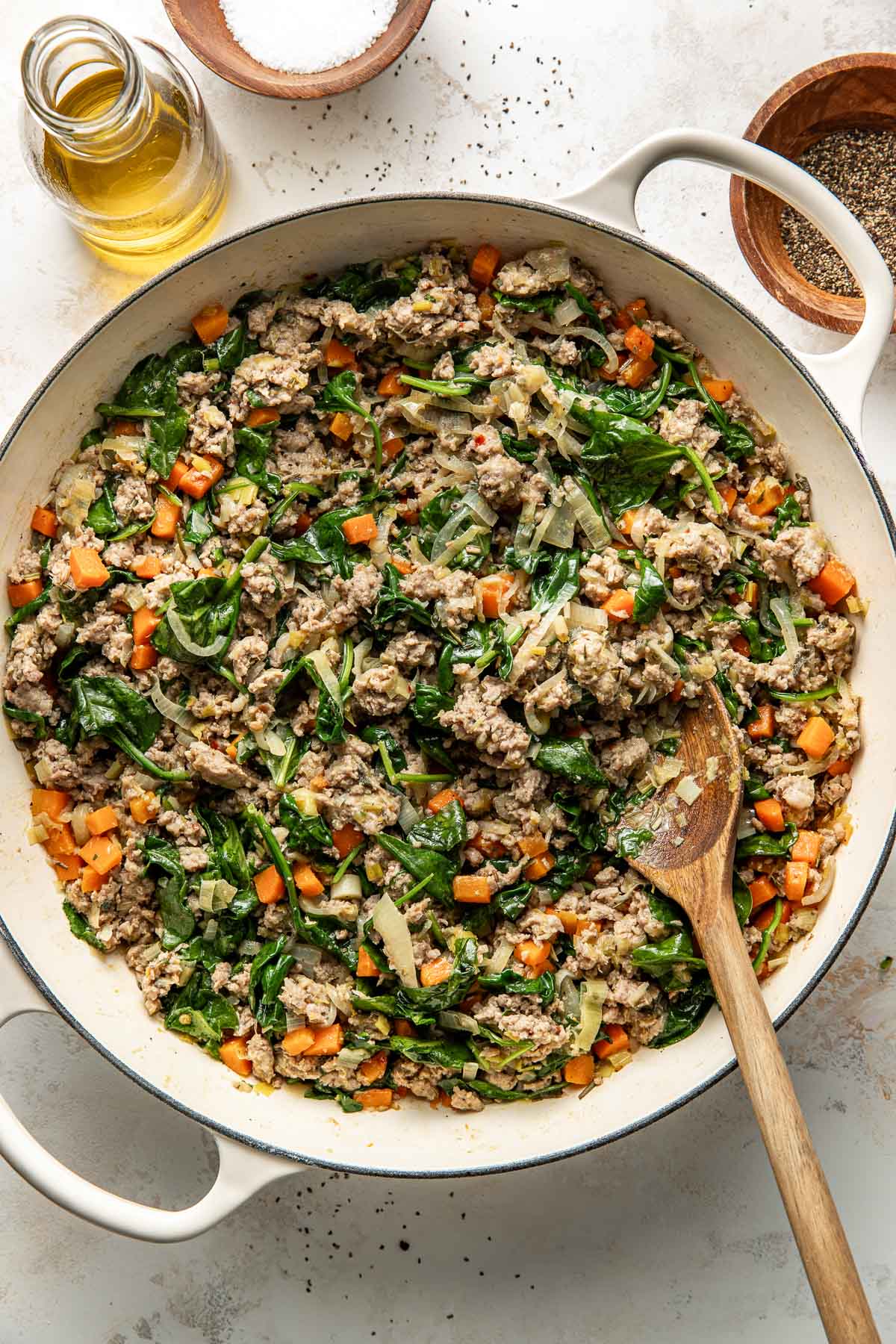 A large white pot filled with a cooked mixture of ground meat, chopped carrots, spinach, onions, and seasonings, with a wooden spoon inside; oil, salt, and pepper containers are beside the pot.